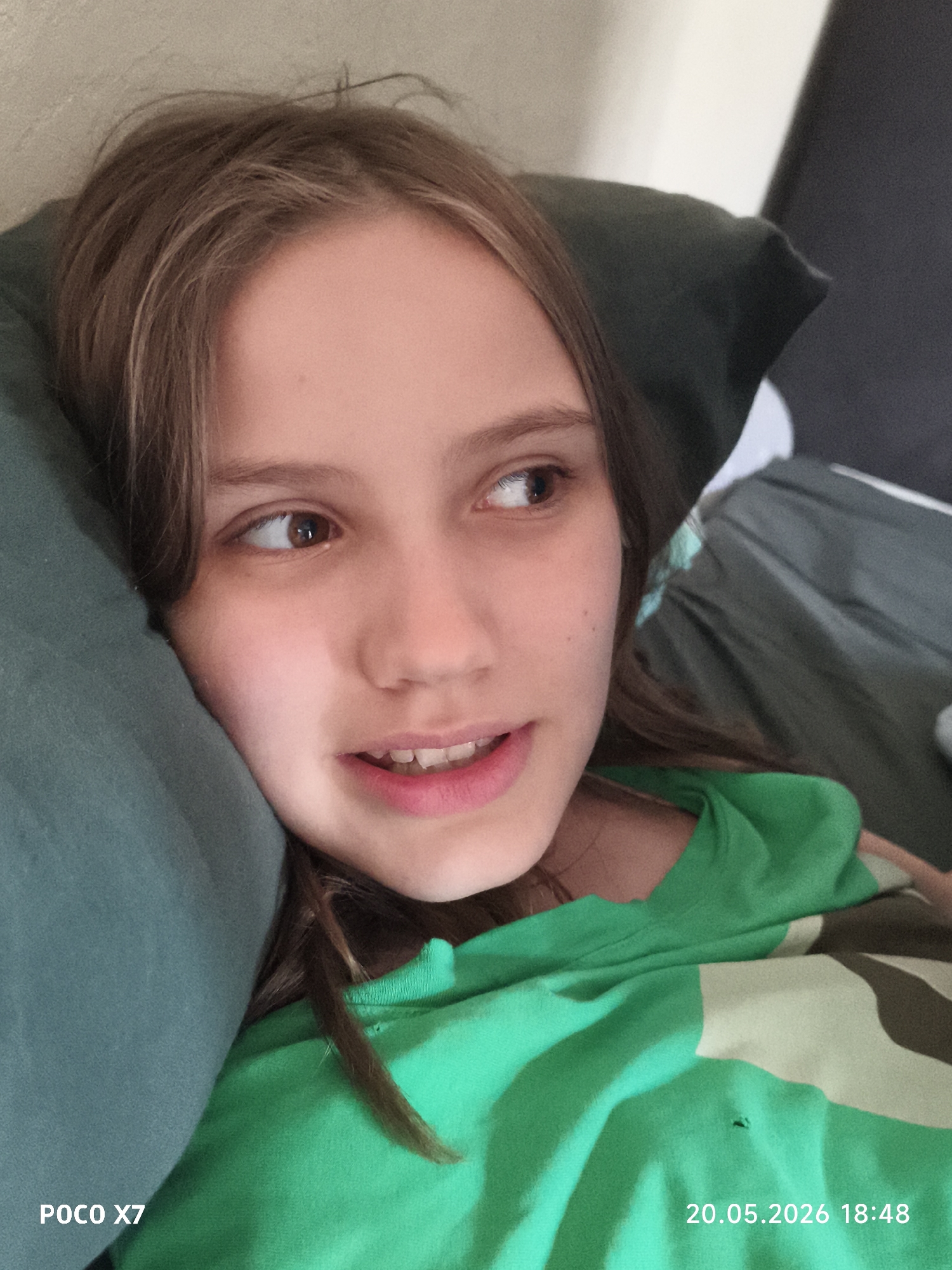 A young person is lying down on a pillow and looking to the side with a slight smile. The pillow is a dark green color. The person has light brown hair and brown eyes. They are wearing a green and camouflage patterned shirt. The bottom left corner of the image has "POCO X7" printed on it, and the bottom right corner has "20.05.2026 18:48". Location is unknown.