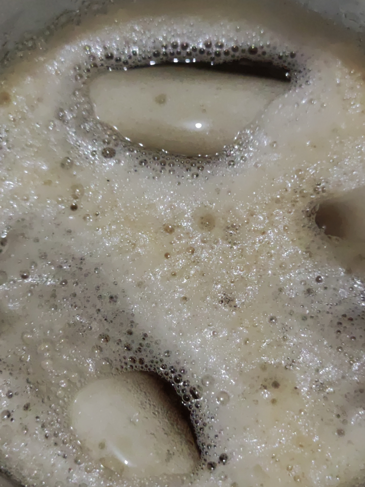 This close-up, top-down image captures the surface of a frothing, light brown liquid in what appears to be a cooking pot or vessel. The liquid is covered extensively by a dense layer of bubbles and foam of various sizes, ranging from tiny specks to larger, iridescent spheres. These bubbles suggest that the liquid is either actively boiling, simmering, or has recently undergone vigorous agitation.
Several irregular, smoother patches of darker liquid are visible beneath the foam, reflecting some light and hinting at the depth of the contents. The overall color palette consists of off-white and various shades of brown, indicating potentially rich ingredients. The texture is highly dynamic due to the numerous bubbles.
The scene appears to be an indoor setting, likely a kitchen, focusing solely on the culinary preparation. Given the location context of Lhokseumawe, Indonesia, this could be a local dish involving ingredients that produce such foam, possibly a broth, a curry, or a sweet concoction. There are no people or discernible text in the image.