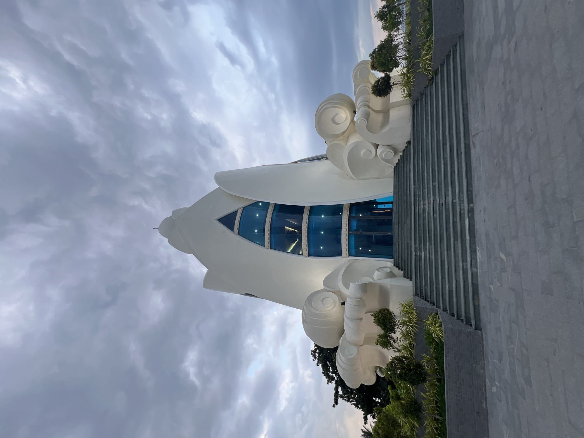 A grand, white, modern architectural structure stands prominently against a dramatic, cloudy sky. The building features a unique, organic design with flowing lines and a tall, pointed roof topped by a small dome. A large, triangular, blue-tinted glass facade is set into the center of the building, with doors visible at its base. Flanking the entrance are sculptural elements resembling swirling waves or clouds, rendered in the same white material as the main structure. A wide, gray stone staircase leads up to the entrance, bordered by lush green landscaping and planters. The ground in front of the staircase is paved with gray stone tiles. Trees and foliage are visible on either side of the structure. The overall impression is one of artistic and contemporary design, likely a religious or cultural monument. This structure is the Long Son Pagoda in Nha Trang, Vietnam.