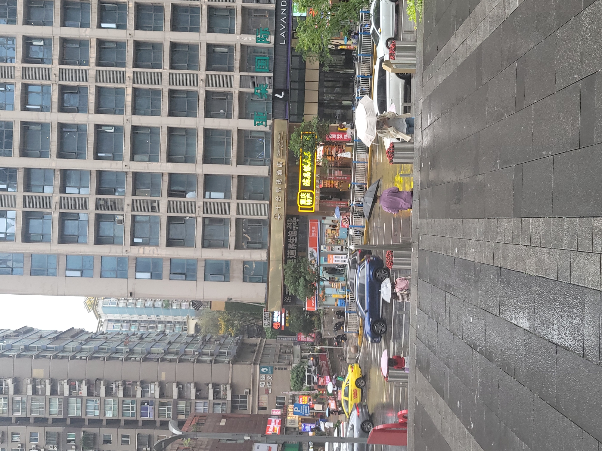 This image depicts a city street scene in Liangjiang, China, during what appears to be a rainy or recently rained-upon day, as indicated by the wet pavement and people holding umbrellas. The time of day is likely daytime, given the general illumination.
The scene is dominated by tall, multi-story buildings that line the street. On the left, a large, modern building features a facade composed of numerous square windows arranged in a grid pattern, with horizontal grilles between some floors. Further back and to the left, another building shows rows of what appear to be air conditioning units protruding from its windows.
At street level, various businesses occupy the ground floors of the buildings. Prominent signage is visible, including "千合智选酒店" (Qianhe Zhituan Hotel) and its English equivalent "Qianhe Zhituan HOTEL," "LAVAND," "圣道养生馆" (Shengdao Health Club/Center), "重庆特产批发" (Chongqing Specialty Wholesale), and a "便利店" (Convenience Store). Other visible Chinese text includes "明德作文" (Mingde Composition), "唐潮," and the number "240." A brand name "EVE" is also seen.
The street itself is wet and features multiple lanes. Several vehicles are present, including a blue SUV, a yellow taxi, and a white car. Metal barriers separate the sidewalk from the road, and small planters with red flowers are placed along the barriers. On the wide, dark-tiled sidewalk, pedestrians are visible, some walking with white, black, or pink umbrellas. One person in a light-colored jacket and jeans holds a white umbrella, while another in a purple top uses a black umbrella, indicating ongoing rain or the aftermath of a shower.