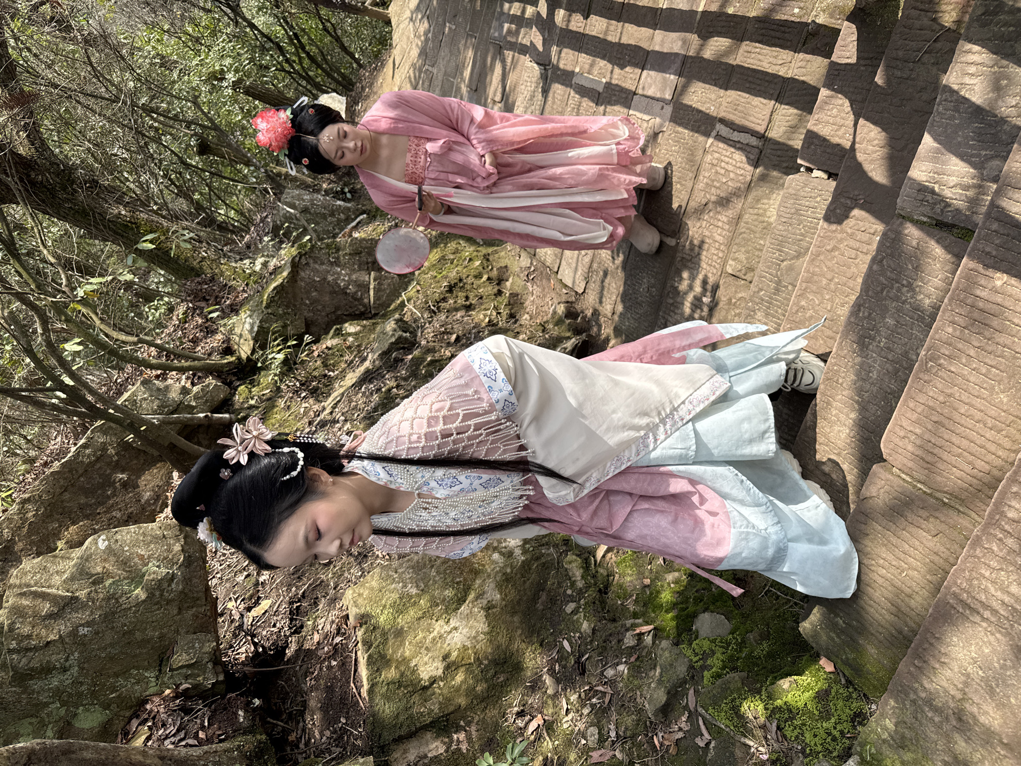 Two East Asian females, dressed in traditional Hanfu, are depicted descending a stone staircase in a natural, forested environment within Wulingyuan, China. The subject in the foreground, positioned left, wears a multi-layered pink, light blue, and cream Hanfu with a pearl-beaded mesh overlay. Her black hair is styled in a high bun adorned with pink and white floral hairpins and pearl strings. She is looking downward as she steps, wearing white athletic shoes. The second subject, in the background on the right, wears a flowing pink Hanfu, featuring a large red floral hair ornament and a gold-colored forehead decoration. She holds a pink translucent fan and a dark object, possibly a mobile phone, in her hands. She stands looking downward on the steps, wearing light-colored flat shoes. The environment consists of rough-hewn stone steps partially illuminated by sunlight, casting prominent shadows. Large moss-covered rocks are visible on the left, and the background is filled with dense trees and undergrowth, indicating a natural, mountainous setting.