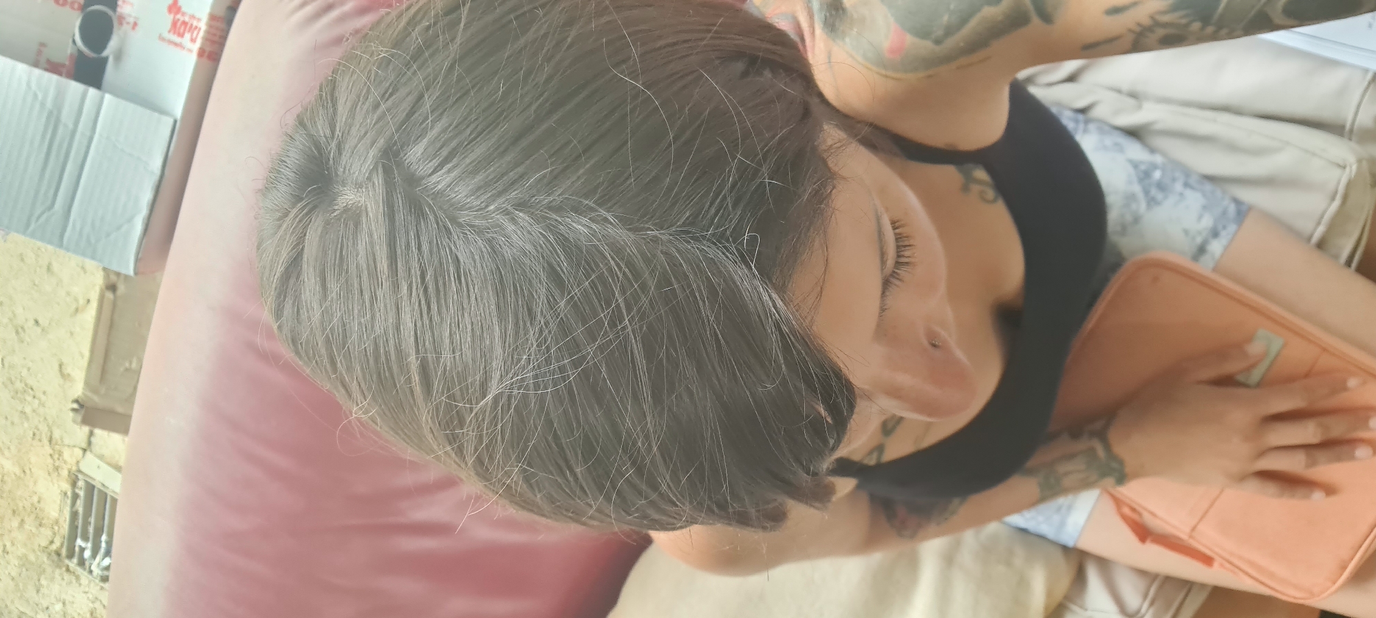A person with tattoos lies on a surface with a pink cushion and tan material. They wear a black top and patterned shorts, holding a peach-colored zippered folder. The setting appears to be outdoors, possibly in the Lachish Regional Council, Israel, though specific landmarks are not visible. A white box is near the person's head.
