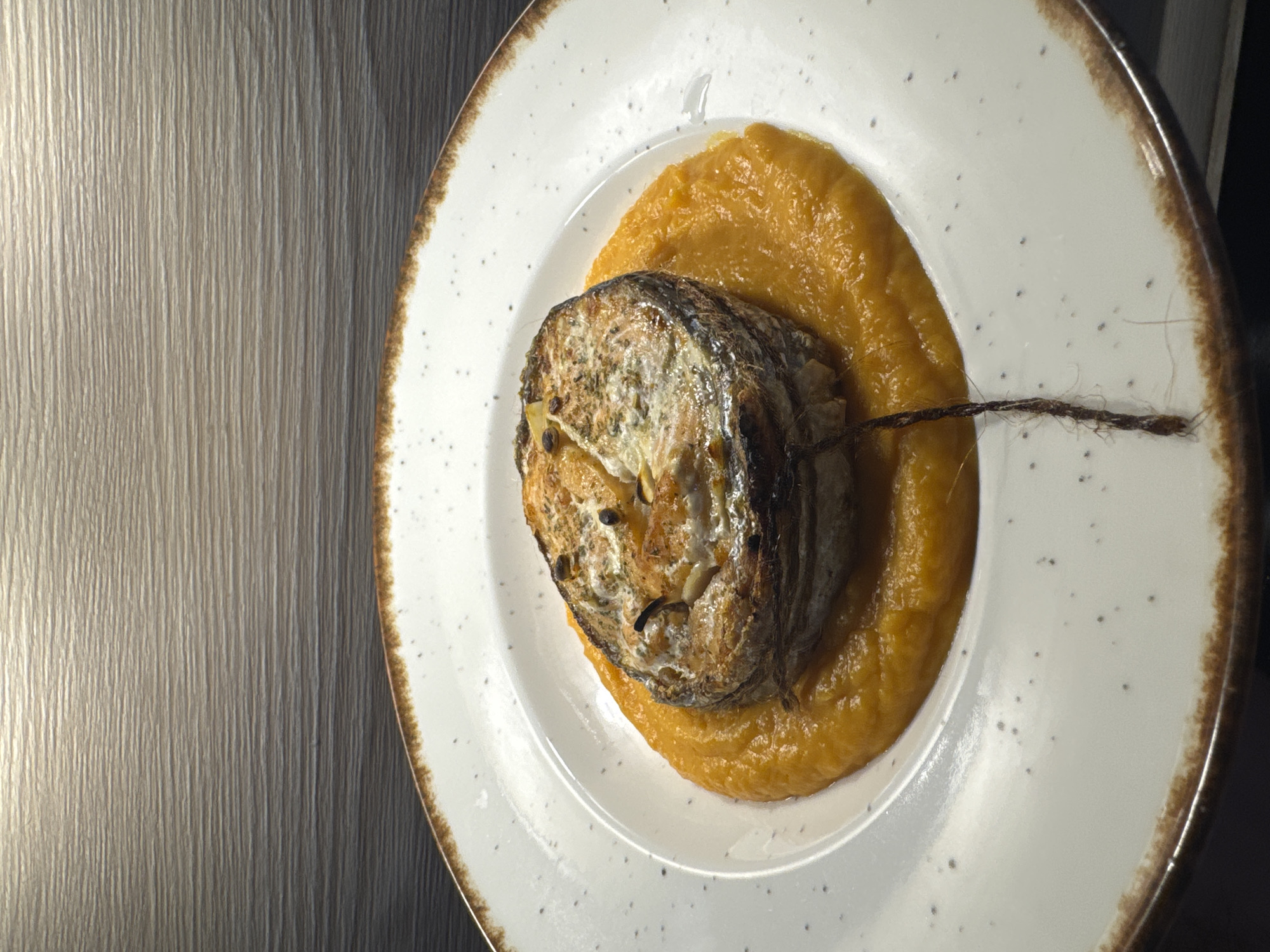 A close-up, overhead shot shows a white plate with a brown rim, speckled with dark dots. Centered on the plate is a portion of vibrant orange purée, upon which sits a thick, circular cut of baked fish. The fish has a golden-brown, crispy exterior, with visible herbs and seasonings. A thin, dark string hangs down from the fish, resting on the purée. The background features a textured, horizontal-lined surface, appearing to be a wooden wall or panel, with a band of light across the top. The lighting is warm, creating a slight sheen on the food and background. The overall scene is a presentation of a prepared dish, likely in a restaurant setting. The location is Unknown, Russia.