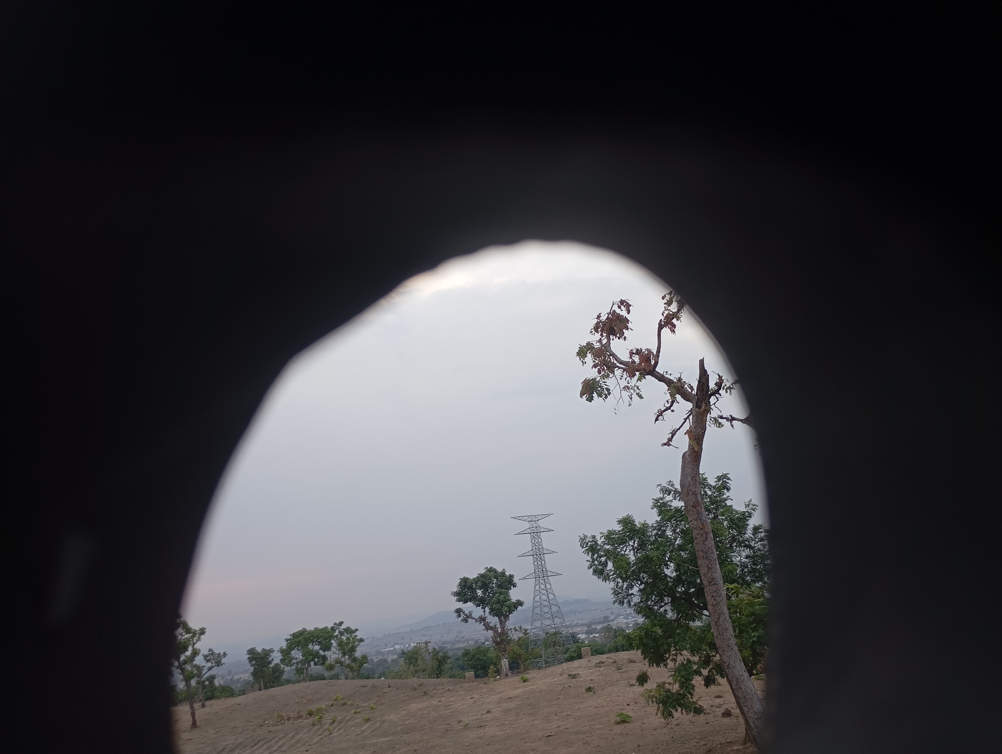The image, potentially taken in Uke, Nigeria, shows a landscape viewed through an irregularly shaped dark opening, possibly a hole. The visible scene includes a bare, dirt-covered terrain with scattered vegetation. In the distance, there's an electrical transmission tower. Several trees are visible. The sky is overcast and pale, indicating either dawn, dusk, or a cloudy day. The foreground is dominated by the dark shape of the opening, obscuring a significant portion of the view.