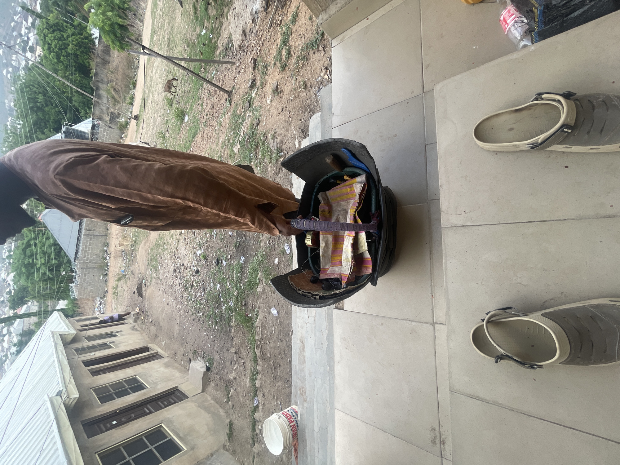 The image, rotated, depicts an outdoor scene in what appears to be a semi-urban or rural setting. In the foreground, on a light-colored tiled floor, are two gray and beige clogs, one of which clearly shows the brand name "CROCS". A white plastic bucket is also visible, with partial text "TOP PUR..." or "TOP PURE" on its side.
To the left, a person, dressed in flowing brown traditional robes, stands beside an open, dark-colored satchel or bag. The bag contains various items, including a striped fabric in shades of yellow, pink, and white. The person's face is not clearly visible.
Beyond the tiled area, the ground transitions to dirt and sparse green patches, with some scattered debris. In the mid-ground, a small, light-brown goat is seen grazing near some wooden poles. Further to the left, a concrete building with multiple dark-framed windows and doors is visible, suggesting ongoing construction or a recently built structure.
The background reveals a hilly landscape dotted with more buildings, indicative of a larger settlement. The lighting suggests it is daytime, possibly overcast, as there are no distinct shadows. The overall setting, combined with the context of Lokoja, Nigeria, points to a common daily scene in the region.