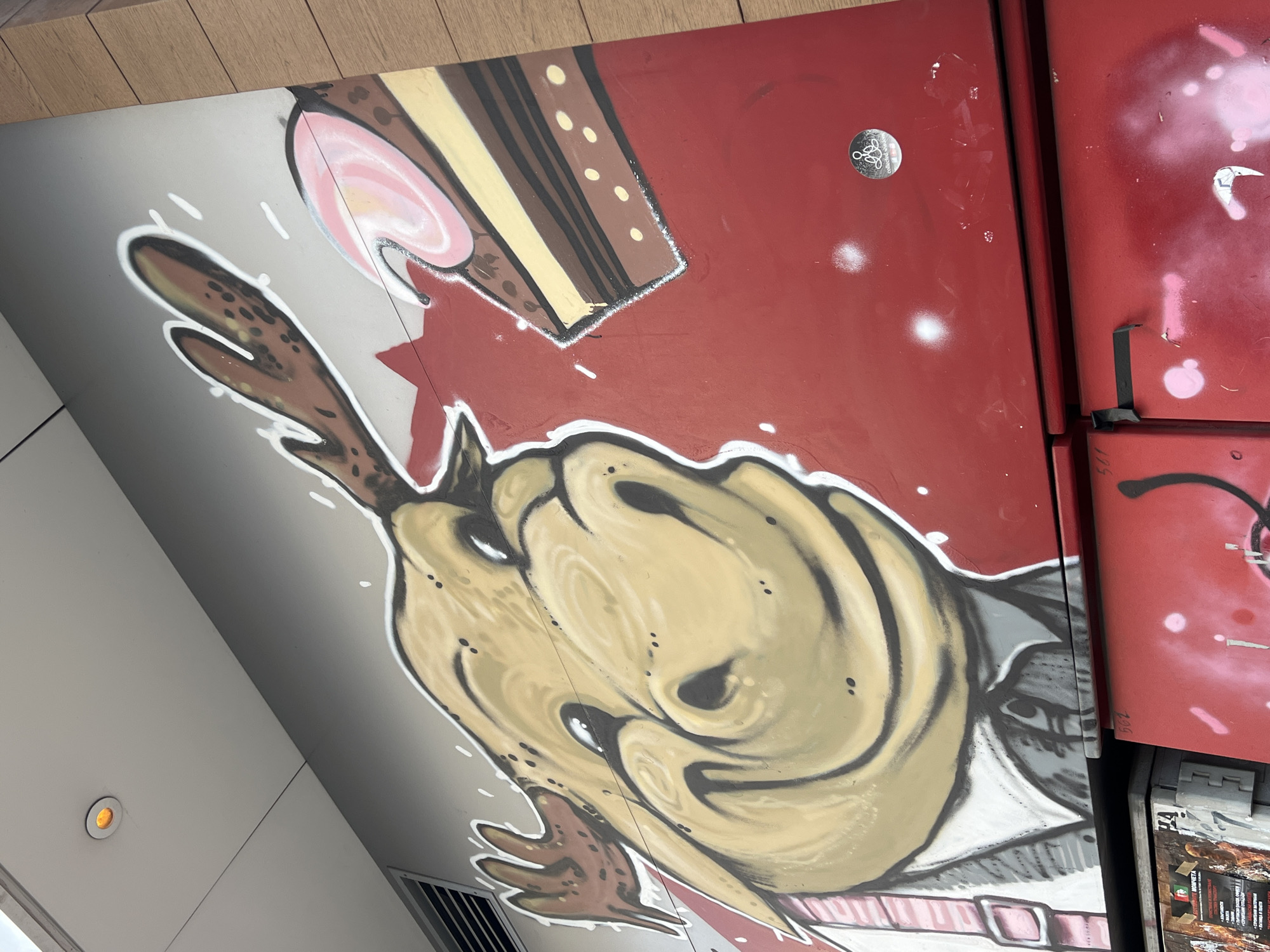 The image shows a graffiti-style mural on a wall or structure in Sofia, Bulgaria. The mural features a cartoonish depiction of a deer-like creature with large, swirling eyes and antlers, wearing what appears to be a suit or attire with a pink belt. Above the creature is an illustration of a slice of cake with pink frosting. The background consists of red and grey surfaces with splatters and other graffiti elements. The setting appears to be an urban environment, as suggested by the metal grid and the adjacent red surfaces with signs of wear. The mural is likely an example of street art or urban art in Sofia.
