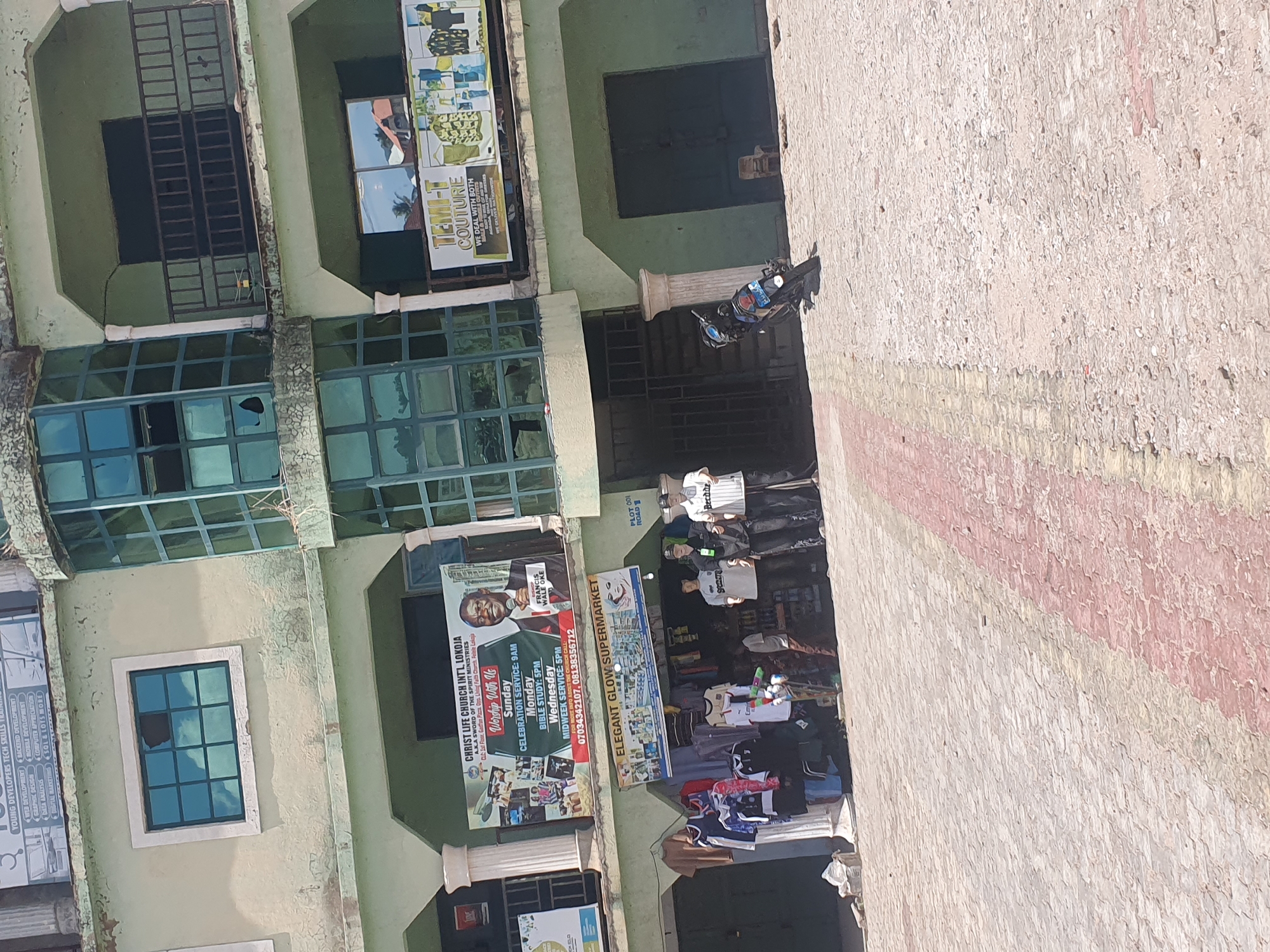 The image depicts a vibrant commercial street scene in Lokoja, Nigeria, under bright daylight. A light-colored, multi-story building with a slightly weathered facade occupies the left side, featuring multiple windows, some with security bars and others appearing cracked or broken.
At the ground level, several businesses are evident. A large banner for "CHRIST LIFE CHURCH INTL, LOKOJA (A.K.A SWORD OF THE SPIRIT MINISTRIES)" advertises "Worship With Us" with specific service times: Sunday Celebration Service at 9 AM, Monday Bible Study at 5 PM, and Wednesday Midweek Service at 5 PM. Contact numbers 07034342107 and 08138356712 are displayed, alongside a portrait of "FRANCIS WALE OKE." Next to this, "ELEGANT GLOW SUPERMARKET" is visible, with several individuals gathered near its entrance, some browsing merchandise displayed outside.
Further down, a sign for "TEMI-T COUTURE" mentions "WE DEAL WITH BOTH" and "WE DELIVER." A partially visible sign advertises "YOUNG DEVELOPERS TECH SKILLS," including "WEB DEVELOPMENT," "GRAPHIC DESIGN," "MOBILE DEVELOPMENT," "DATA ANALYSIS," "BACKEND DEVELOPMENT," and "DIGITAL MARKETING."
A motorcycle is parked near a decorative pillar on the right. The adjacent building or wall on the far right has a textured, light-colored surface with distinct red and yellow striped sections. The overall scene portrays a bustling commercial area.