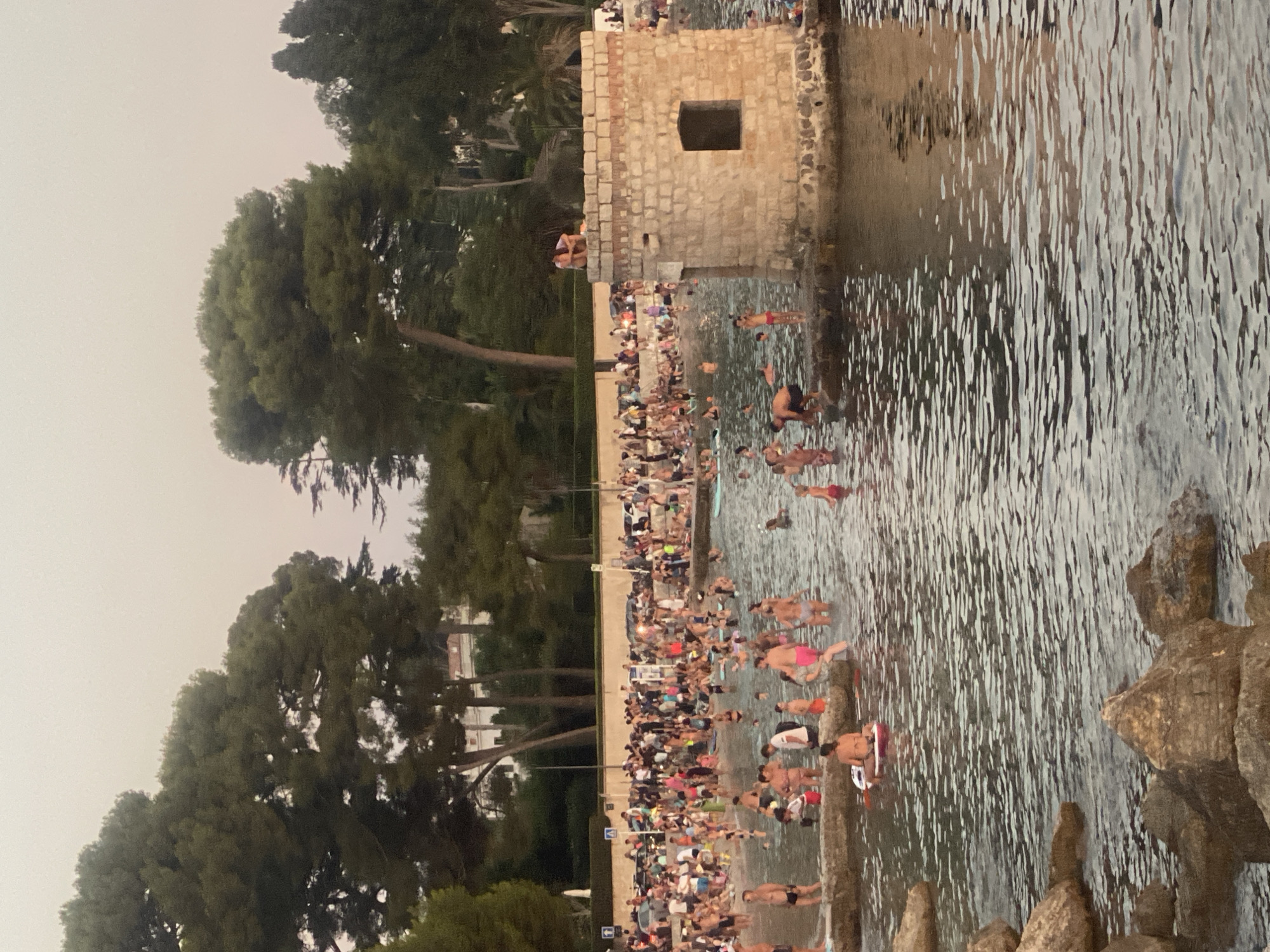The image depicts a crowded beach and waterfront scene in Antibes, France, likely during the summer evening. In the foreground, rocks are visible in the water, with the surface rippling. Further out, many people are wading in the water, swimming, and standing on the shore. Some are on paddleboards. A stone structure, possibly a small tower or fort, juts out into the water on the right side of the image. Two people are perched on top of this structure. Behind the beach, a dense line of trees, primarily pine trees, forms a backdrop, with buildings visible beyond the trees. Cars are parked along the road behind the beach. The sky is a light, hazy orange, suggesting sunset or possibly atmospheric conditions. The overall impression is one of a lively and popular recreational area.