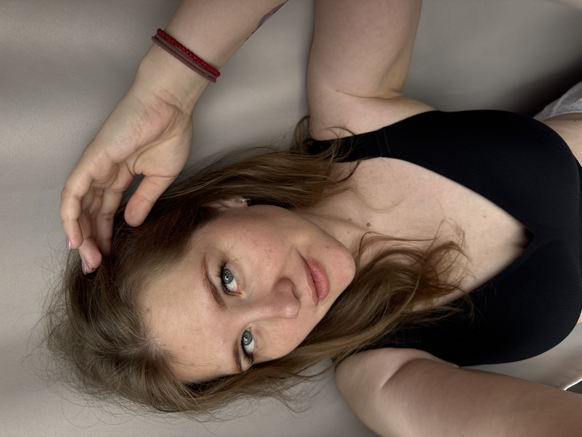A woman with light brown hair and blue eyes is lying on a light beige surface, possibly fabric or a bed covering. She is wearing a black top. Her left hand is raised and placed near her head. A red bracelet adorns her left wrist. The image appears to be taken from a high angle, looking down at the subject. There is no indication of the location being Gornyi (Dvurechenskoe S/P), Russia.