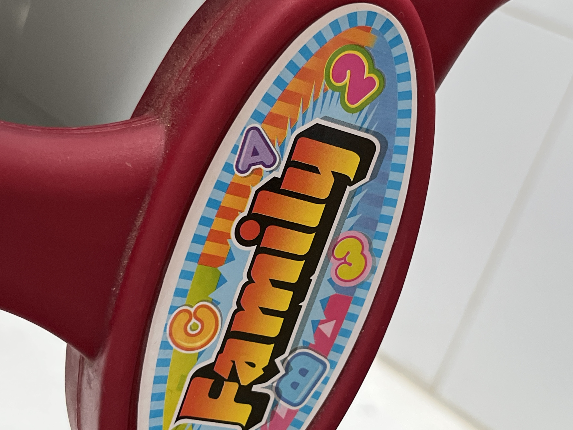 The image shows a close-up of a red plastic object with a colorful sticker on it. The sticker features the word "Family" in a cartoonish font, with each letter a different color and stylized with a black outline. The background of the sticker has various shapes and the letters "A" and "B" as well as the numbers "2" and "3" scattered around. The plastic object has a slightly dusty or worn appearance. The background is blurry and appears to be a white or light-colored surface. No specific information is present to suggest a connection to Lhokseumawe, Indonesia.