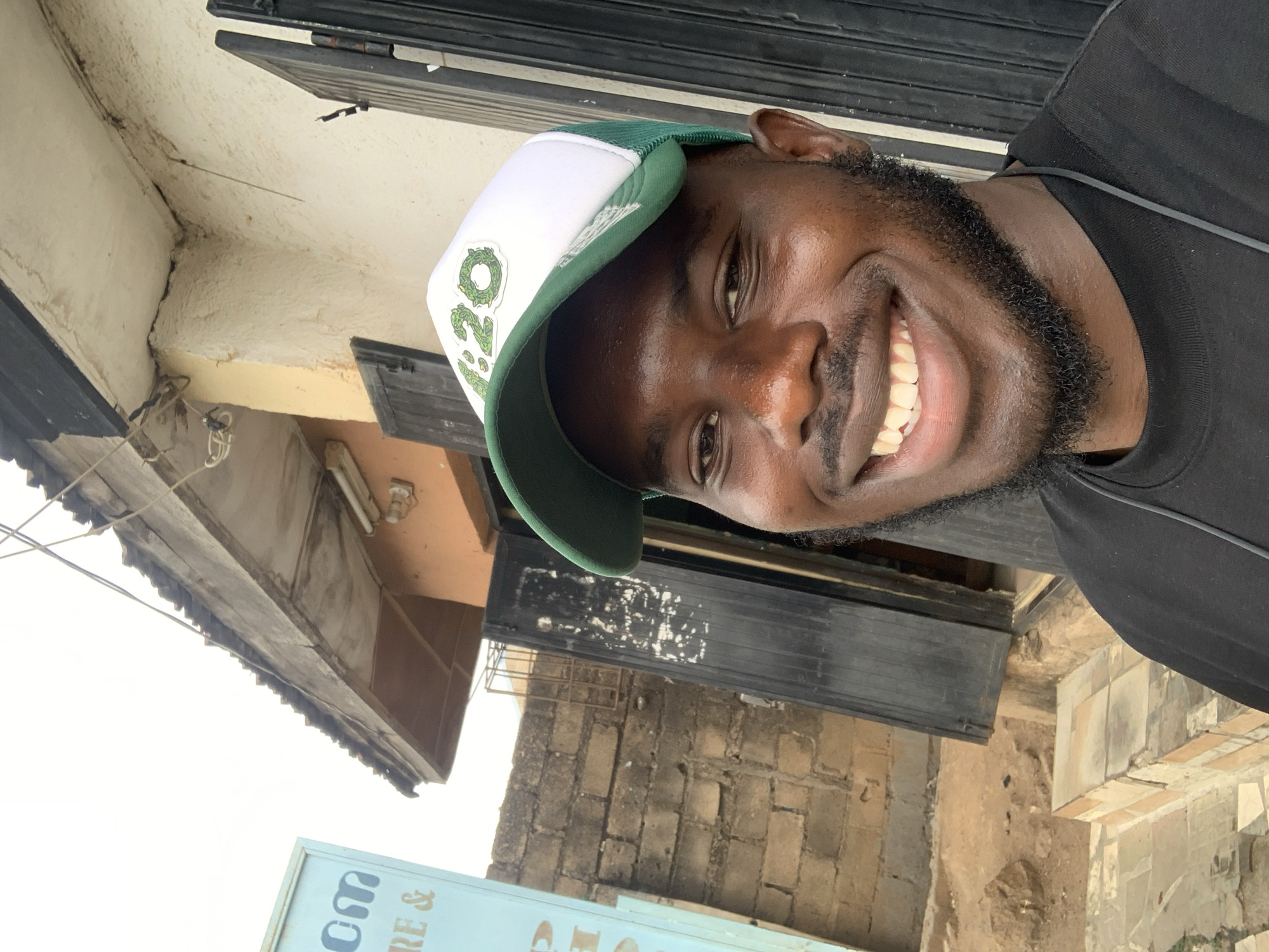 A close-up, brightly lit outdoor portrait features a smiling young Black man looking directly at the camera. He wears a black t-shirt and a white and green baseball cap with a green brim and mesh back. The cap has a prominent green "420" logo, possibly stylized with a leaf design, on its front. A black earbud wire is visible around his neck.
The man has a beard and mustache and is smiling broadly, revealing his teeth, conveying a cheerful and friendly expression. His skin is dark-toned and appears to have a natural sheen.
The background elements suggest an urban or semi-urban setting in Jos, Nigeria. Above the man, there is a light-colored building overhang or ceiling, showing some cracks and a textured finish. Dark, slatted window shutters are visible behind him. Further back, a brick wall can be seen, indicating surrounding structures. In the bottom part of the image, a light blue sign is partially visible, showing text fragments like "OM" and "RE &". The clear, bright lighting indicates it is daytime, likely mid-morning or afternoon.