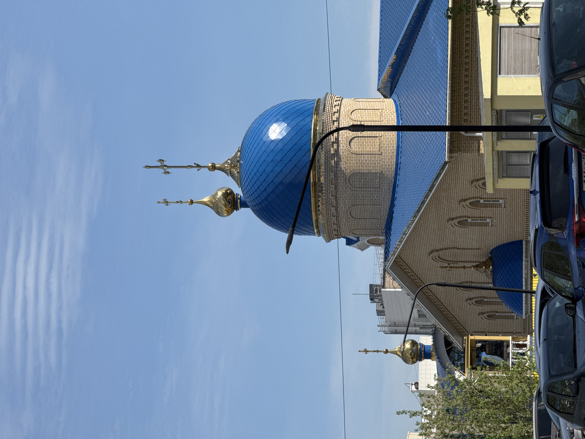 A low-angle, medium shot depicts the Orthodox Cathedral of St. Michael in Izhevsk, Russia, against a clear blue sky. The church has a striking blue dome with gold accents, topped with an Orthodox cross. The dome is tiled and reflects sunlight. The building's facade is beige brick, with decorative arched windows. A black street lamp is in the foreground. A building with yellow walls and closed blinds, along with parked cars, suggests an urban environment. The overall composition is partially rotated.