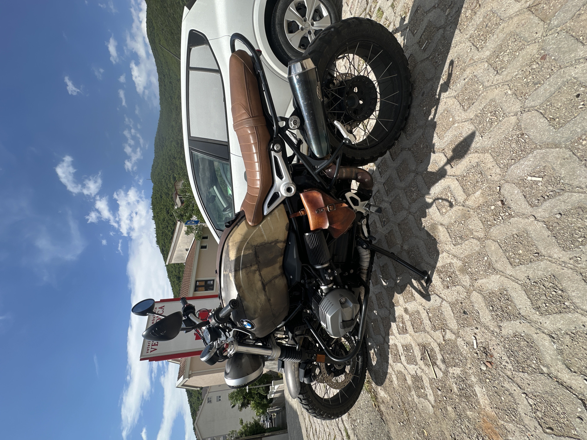 The image shows a BMW R nineT Scrambler motorcycle parked on a patterned concrete surface. The motorcycle has a custom-painted tank, a brown leather seat, and a side-mounted leather bag. It is parked next to a white car. In the background are buildings and mountains. A sign for "Veterinarska Ambulanta Veca" is visible. The location is Budva, Montenegro, based on the sign's name. The sky is blue with scattered clouds.