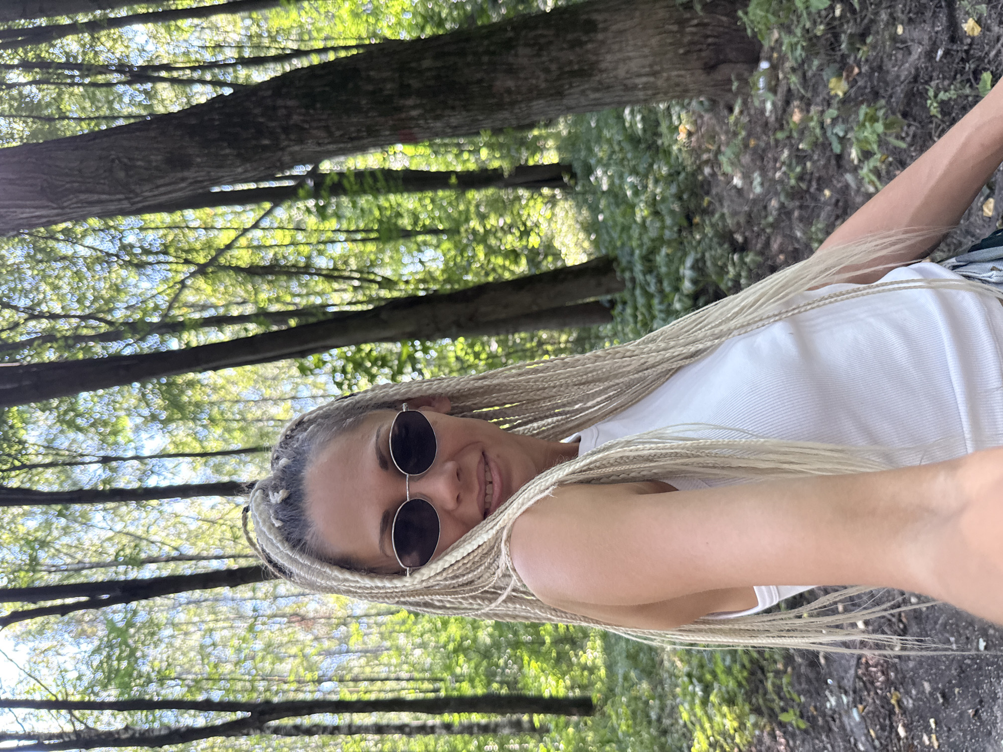 A woman with blonde braided hair and sunglasses is taking a selfie in a wooded area. She is wearing a white tank top. The background is filled with trees and greenery, suggesting a park or forest setting. The lighting indicates it is daytime, likely sunny, with dappled light filtering through the leaves. The ground is covered with dirt, fallen leaves, and some patches of green vegetation.