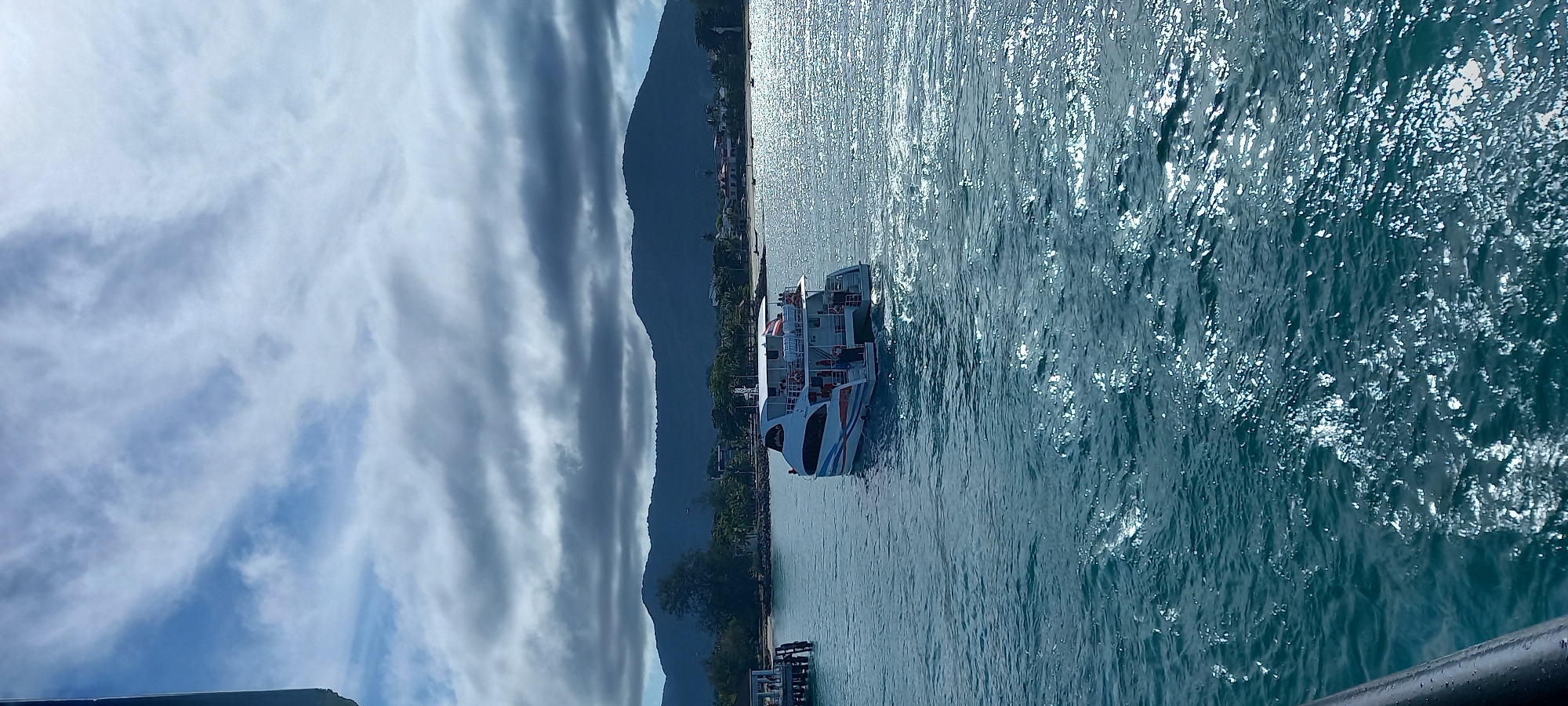 The image shows a passenger ferry docked at a pier in ตำบลเกาะพะงัน, Thailand. The ferry is white with blue and orange accents. Passengers are visible on the ferry's decks. The water is sparkling blue, reflecting sunlight. A mountain range rises in the background. The sky is partly cloudy, with streaks of clouds. A black railing is visible in the foreground. The location is near the coastline, with vegetation and buildings visible along the shore.