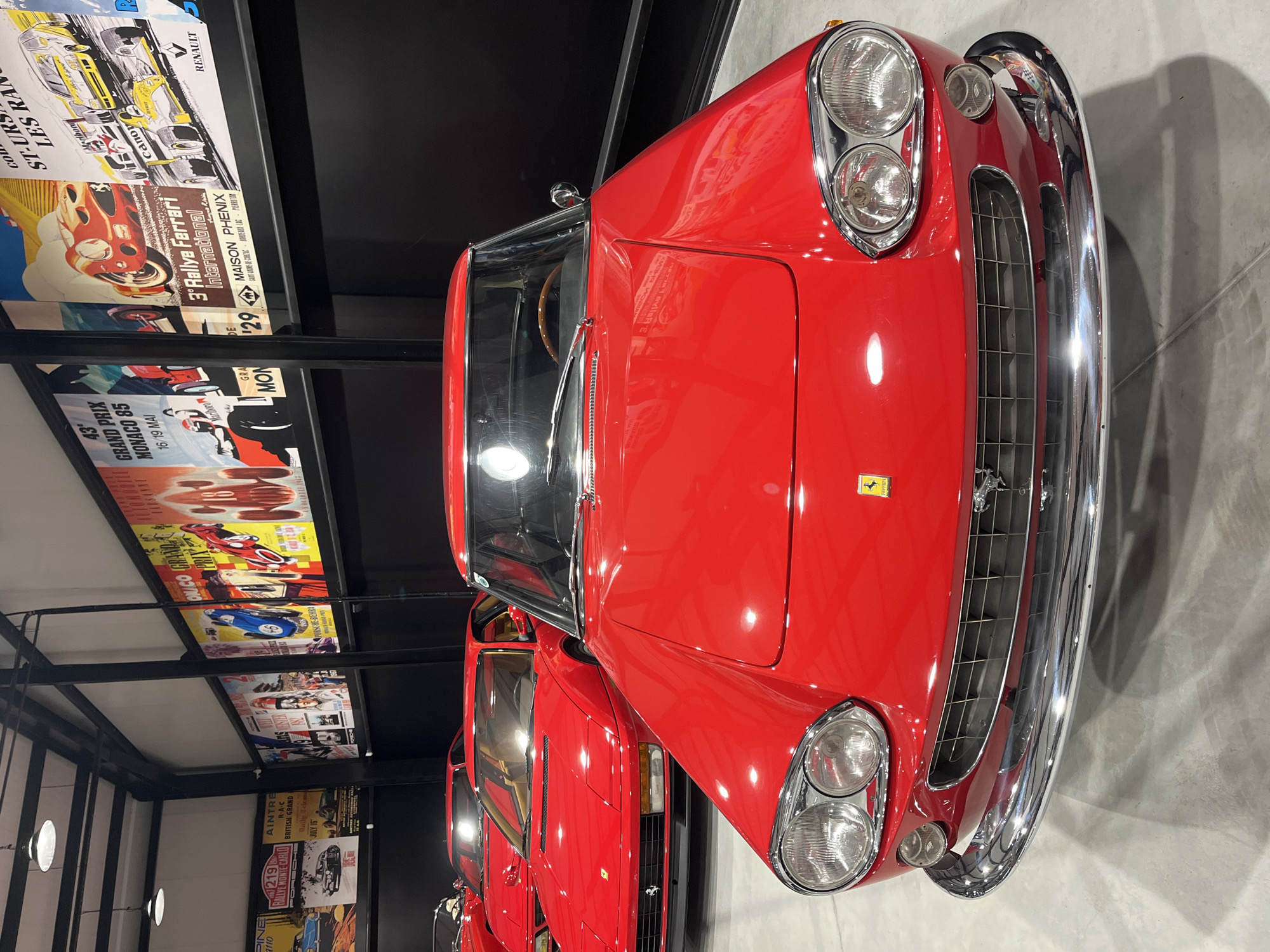 A red classic Ferrari automobile is in focus in this indoor scene. The car is positioned on the right, its hood facing forward, and its shiny chrome bumper is visible. Several vintage motorsport posters adorn the wall on the left, displaying race events and sponsors, creating a backdrop of automobile history. The location and specific actions of people are not identifiable from the given information. The specific location is a car showroom. The location in the city of Kapatovo, Bulgaria, cannot be determined from the image.