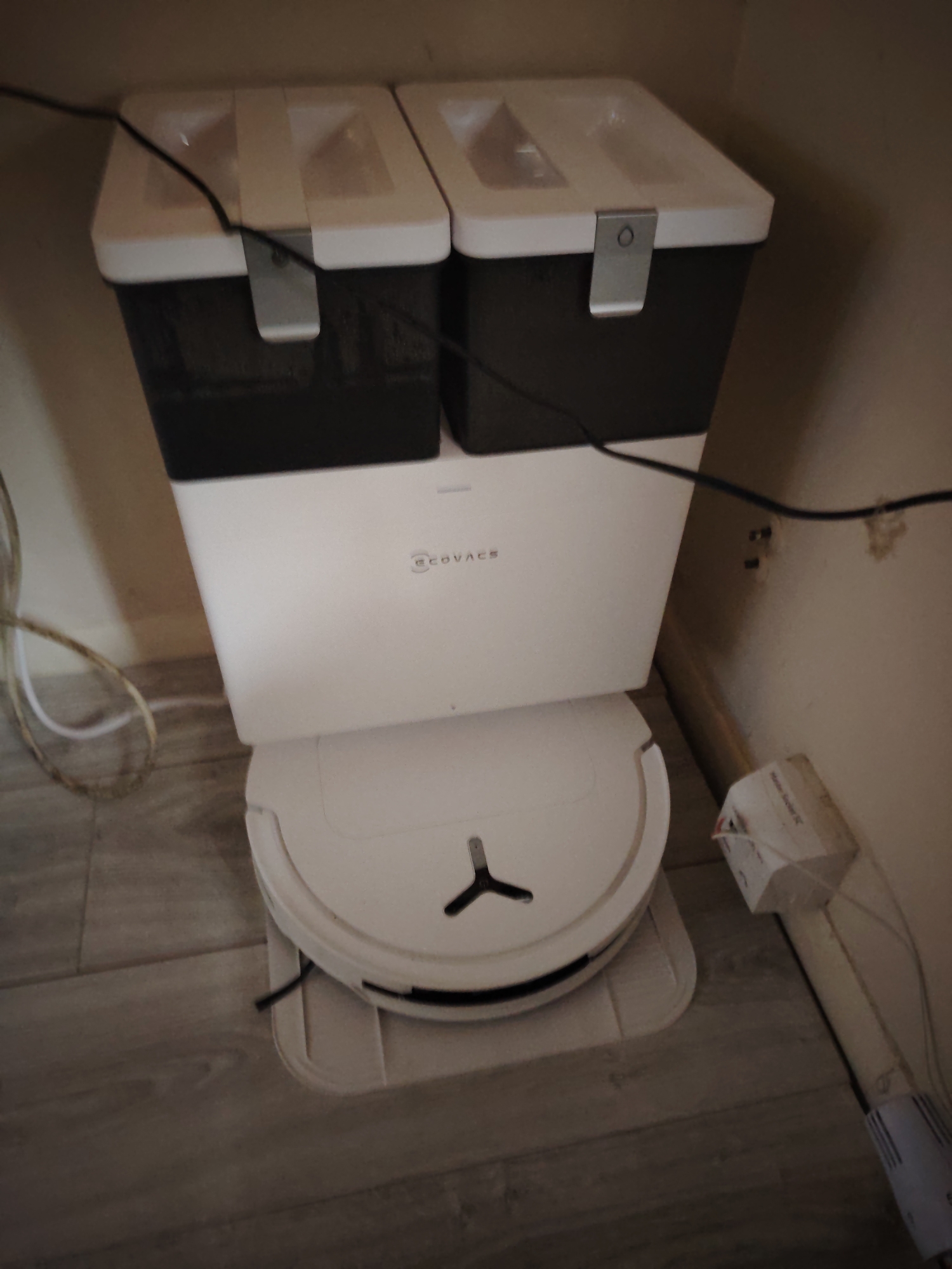 The scene depicts an indoor environment in Huntingdonshire, United Kingdom, featuring a white ECOVACS robot vacuum cleaner charging station and an associated robot vacuum. The charging station consists of a white rectangular base unit with the "ECOVACS" logo prominently displayed on its front face. Stacked atop this base are two dark grey or black rectangular bins, each with a white, semi-translucent lid secured by a metallic, U-shaped latch with a circular perforation.
The robot vacuum, circular and white with a black edge, rests on a slightly elevated white docking mat or plate at the base of the charging station. A black three-pronged brush or sensor is visible within a central opening on the vacuum's top surface.
The entire setup is positioned against a light beige or off-white wall in a corner. The floor is covered with light wood-effect laminate planks. Several cables are present: a black electrical cable runs from behind the charging station, draped over the top of the two bins, and down the right side. On the left, a light-colored coiled cable lies on the floor. On the right wall, a small, white, rectangular electrical box (possibly a power adapter or junction box) is mounted near the floor, with fine wires extending from it. Below this box, another white cylindrical electrical component is partially visible, connected by a cable descending towards the floor. The ambient lighting appears dim. No people are visible.