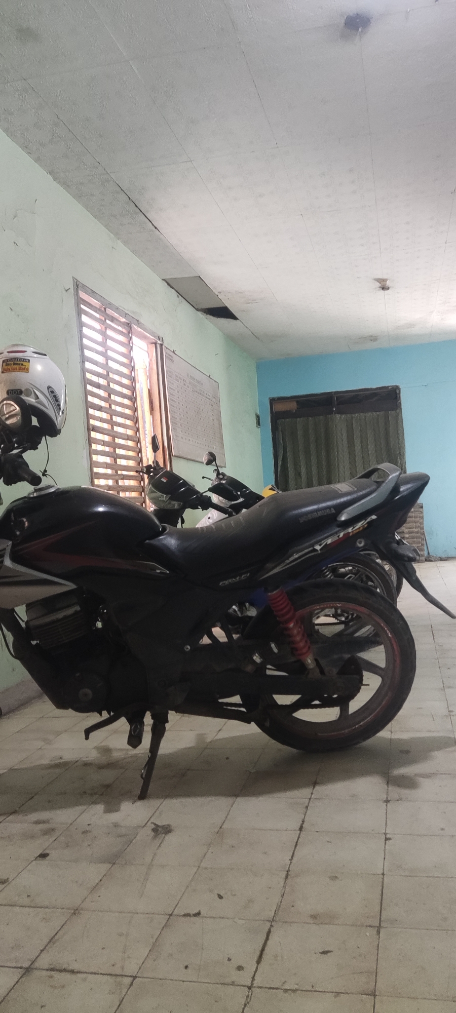 In a room in Gebang, Indonesia, two motorcycles are parked on a tiled floor. The motorcycle in the foreground is black with red and white accents. A white helmet sits on its handlebars. The room has light green walls and a light blue wall in the background. The ceiling appears to be made of acoustic tiles, some of which are damaged. A window with horizontal blinds is visible on the left side of the image. A whiteboard with text and a grid is mounted on the wall to the right of the window. The environment appears to be a storage area or a common space within a building.