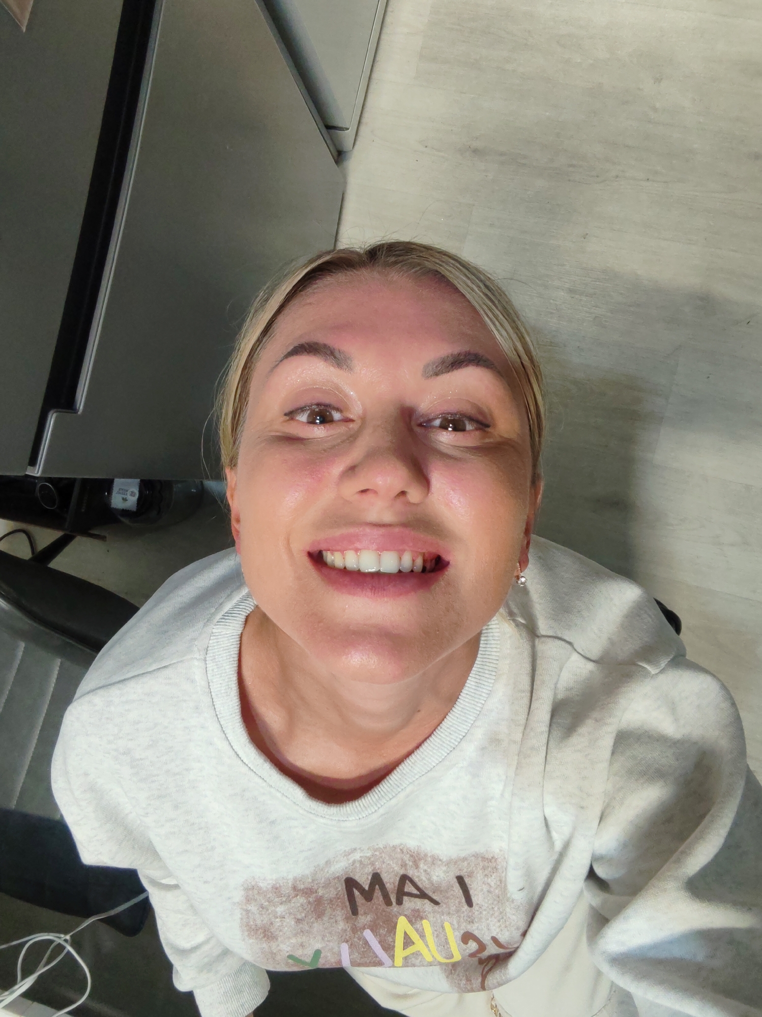 A woman with blonde hair and makeup smiles into the camera in an indoor setting, possibly a home. She is wearing a light-colored sweatshirt with a graphic design on the front. Behind her, a refrigerator and a wooden floor are visible. There is no indication of the specific location being Komsomolsk-on-Amur, Russia.