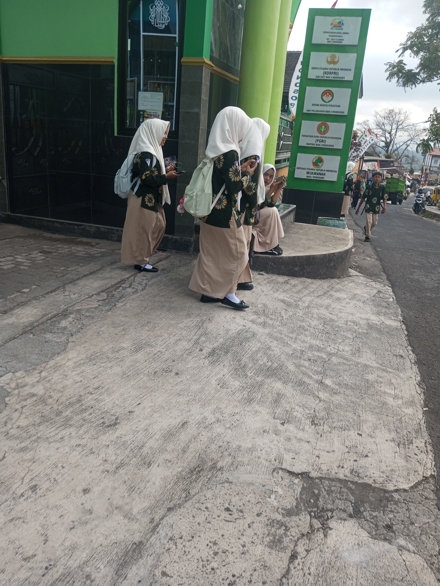 In the city of Krasak, Indonesia, a group of young females wearing hijabs and school uniforms stand on a concrete sidewalk. The uniforms consist of a dark green, patterned top and a tan skirt. Some are carrying light-colored backpacks. One girl is looking at her phone, and another appears to be talking to her. To the right, a young male in a similar uniform walks down the street. In the background, there is a green building with black tiled walls, a white sign with several panels listing organizations, and a street with various vehicles, including a truck and motorcycles. The sidewalk shows signs of wear and tear, with cracks and uneven surfaces.