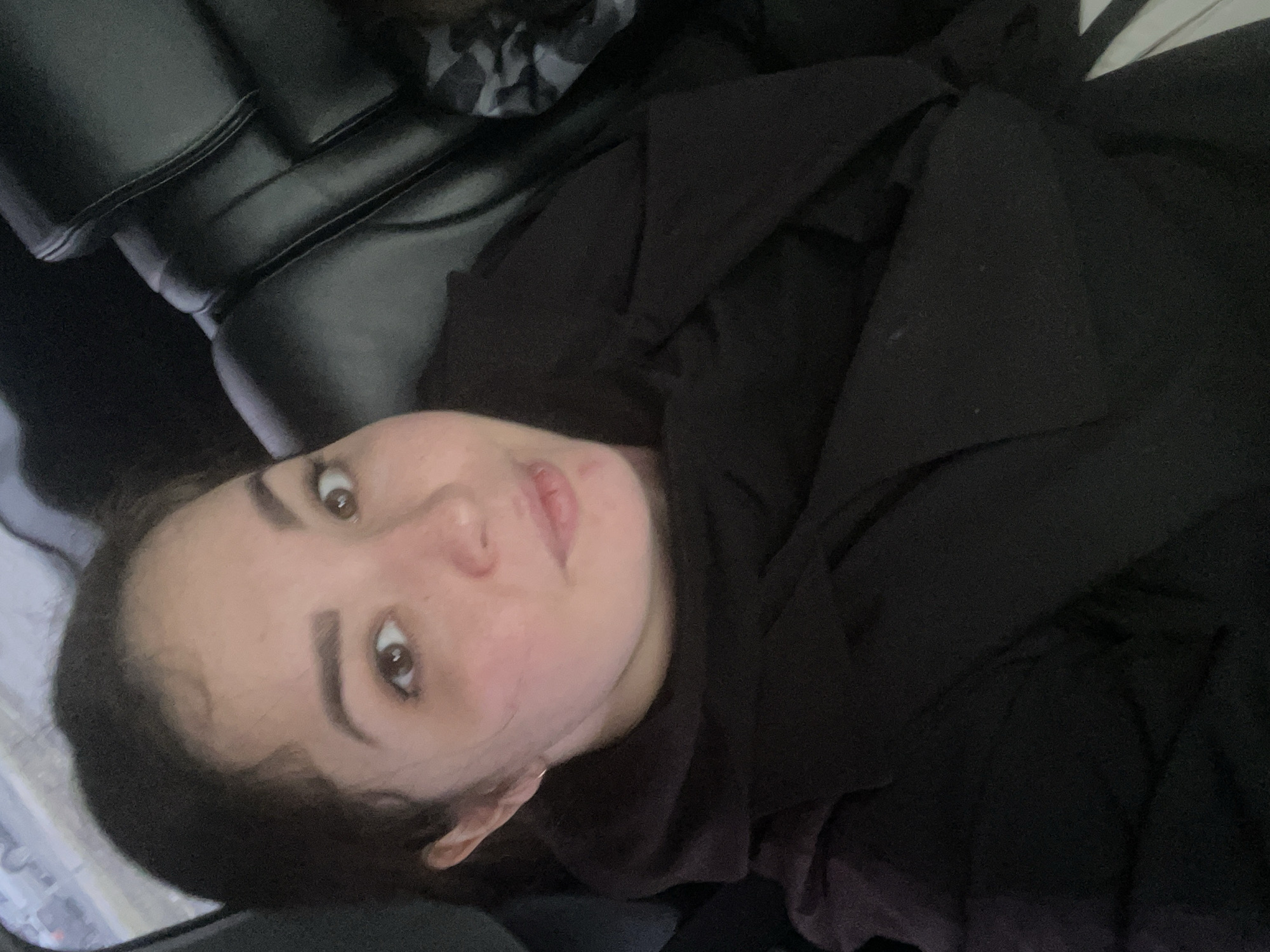 An image captures a young woman with dark hair and brown eyes, looking directly at the camera with a slightly surprised or curious expression and parted lips. Her head is tilted, suggesting she is reclining or lying down, potentially on a bed or sofa. She is dressed in a dark, likely black, hooded top or jacket that covers her body. The background on the upper left features a dark, textured surface, possibly leather upholstery of a piece of furniture. Other areas of the background show soft, dark fabric, hinting at bedding or blankets. The overall lighting is dim, indicating an indoor setting with low ambient light, perhaps during the evening or night. No specific location cues for Izhevsk, Russia are visible in the image itself, nor is any text. The photograph appears to be a casual, unposed shot.