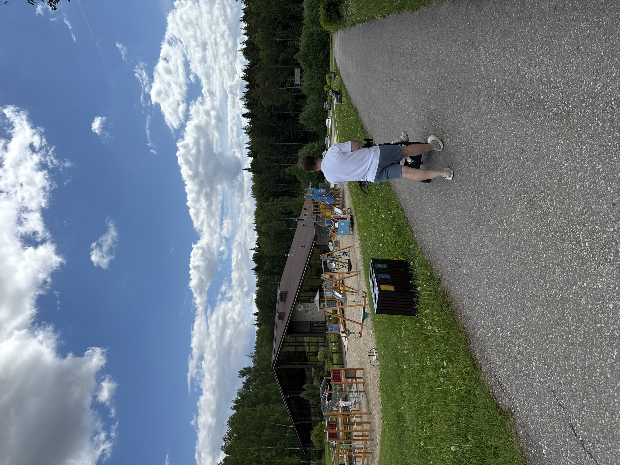 This image captures an outdoor recreational area in Dobrograd, Russia, under a bright, partly cloudy sky. A person, viewed from the back, walks along a paved asphalt path that curves gently. The individual is dressed in a white t-shirt, dark shorts, and light-colored shoes.
To the left of the path, a stretch of green grass dotted with dandelions precedes a modern building with a dark brown roof and expansive glass windows. Through these windows, the Cyrillic text "ДОБРОГРАД" is visible, directly referencing the location context. In front of the building is a large, well-equipped children's playground featuring multiple wooden and metal structures, including slides, swings, and various climbing elements.
Beyond the building and playground, a dense, dark green forest forms a natural backdrop. Adjacent to the path on the right side, a black trash bin with distinct recycling symbols (for bottles, paper, and general waste) is visible.
The sky is a vibrant blue, adorned with numerous fluffy white cumulus clouds, indicating fair weather, likely during late morning or early afternoon. The overall scene suggests a pleasant and well-maintained environment ideal for leisure, walks, and family activities.