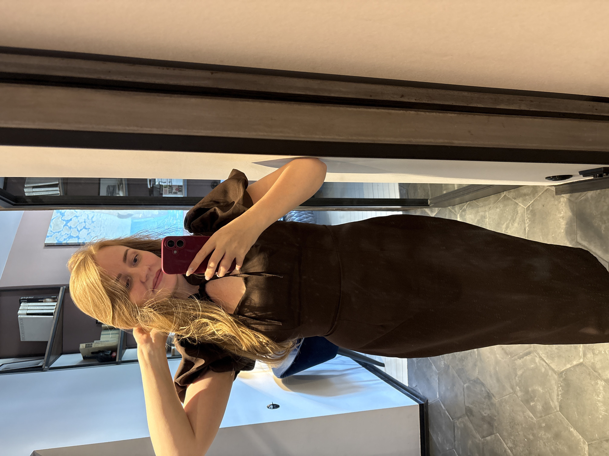 A woman with long blonde hair poses for a selfie in a mirror. She is wearing a brown dress with puffed sleeves and holding a red phone. The background includes a shelf with books and other items, a blue and white painting, a white cabinet, gray hexagonal floor tiles, and a window with a dark frame. The location is likely indoors, possibly in Saint Petersburg, Russia, based on the architectural style and design elements, though without further context or landmarks, this cannot be confirmed definitively.