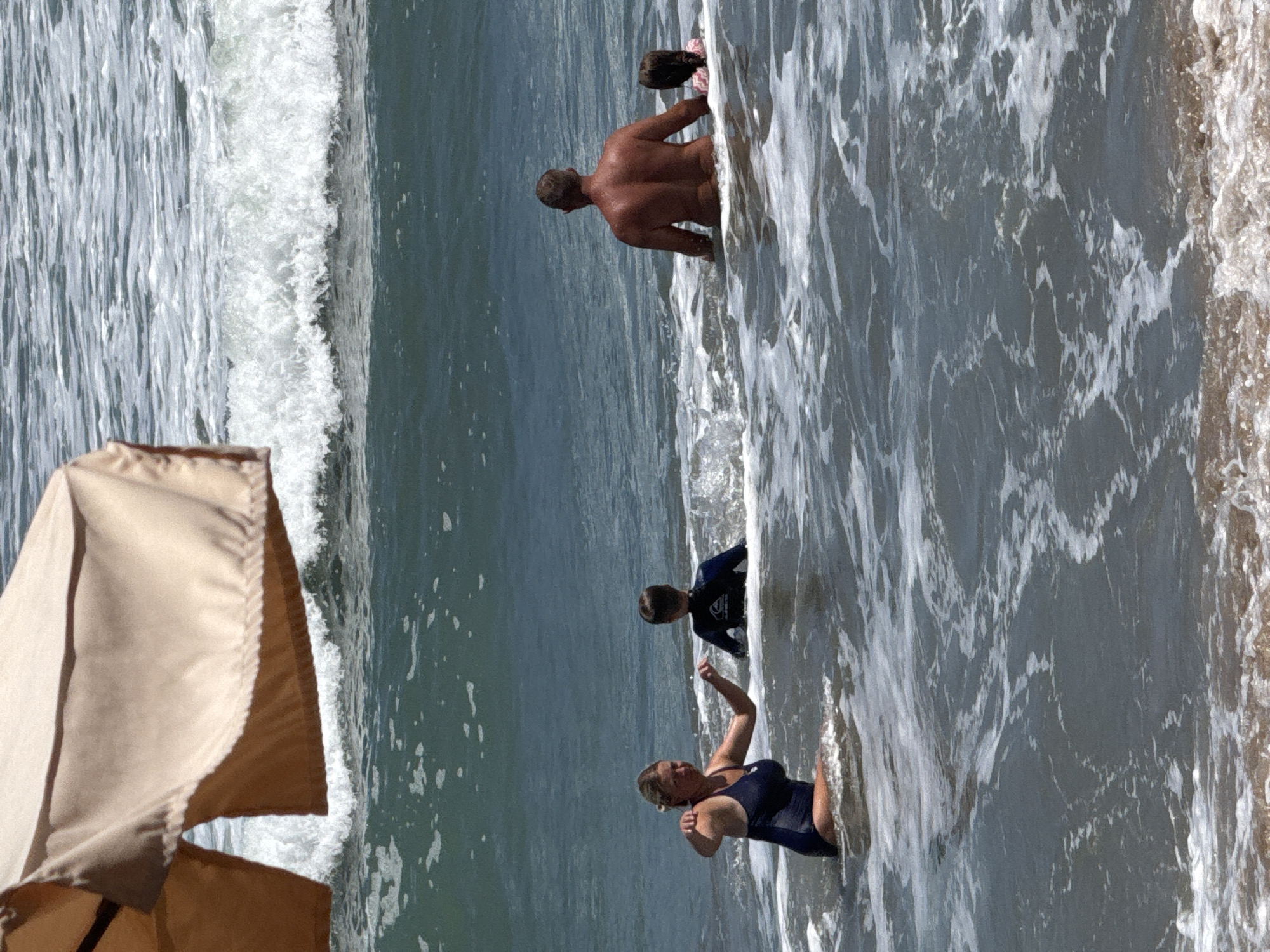 The media file shows a group of people in the ocean at Kuta, Indonesia. The scene is set in shallow, wavy water near the shore. A man, seen from behind and nude, is partially submerged in the water, facing away from the viewer. A child, whose hair is visible, is partially in front of him. Further in the distance, two more people are in the water. One, likely a child, is wearing a dark wetsuit and is looking towards the viewer. The other, a woman in a dark blue swimsuit, is in the foreground, facing towards the viewer, and appears to be swimming or wading. The water is a mix of dark blue and white foam from the breaking waves. The sun appears to be bright, casting shadows and highlighting the water's movement. The presence of a beach umbrella in the foreground suggests a sunny day at the beach. The overall mood seems to be relaxed and recreational, indicative of a day spent at the beach. There is no visible text in the media file.