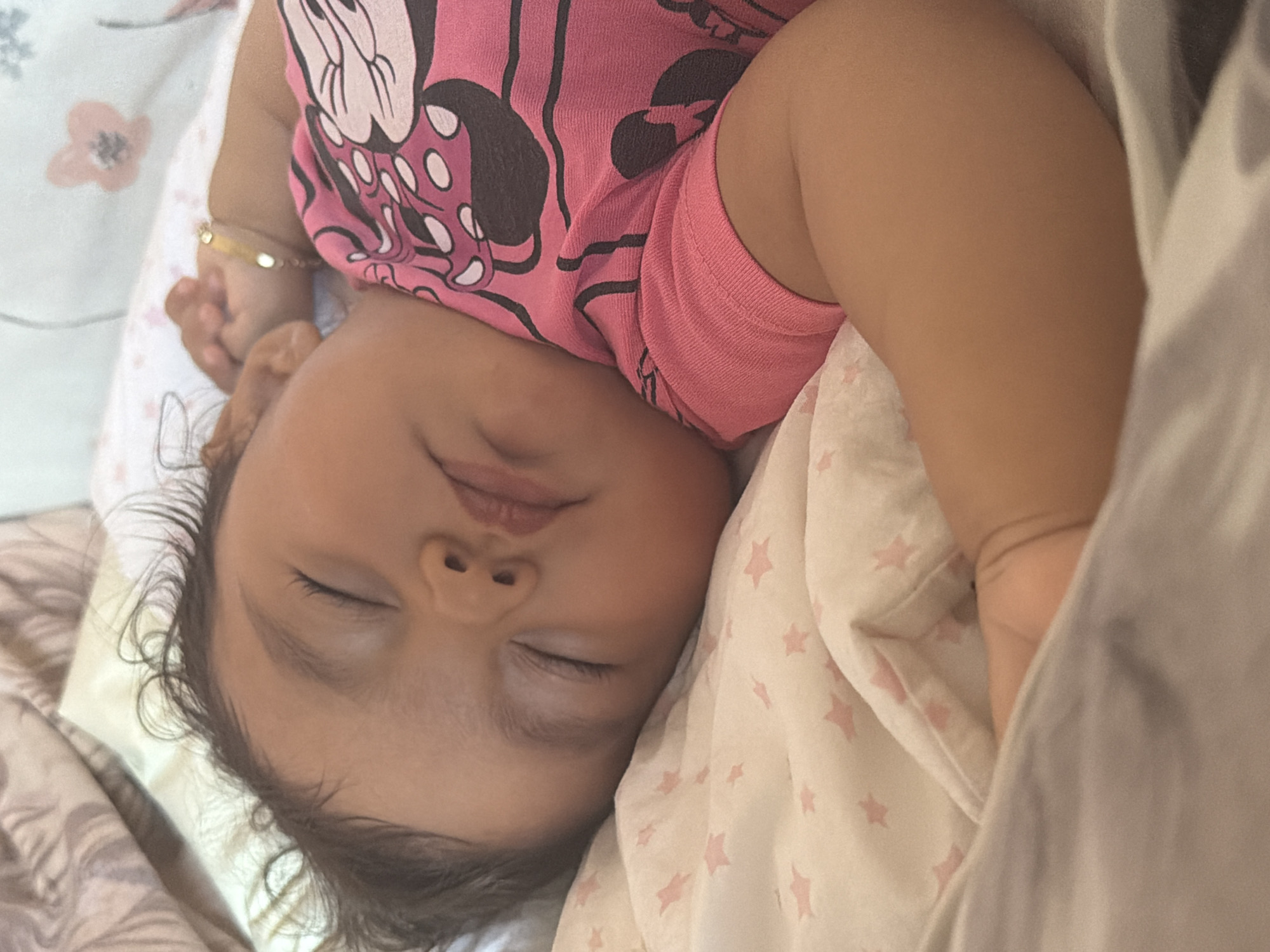 A close-up, high-angle shot shows a baby sleeping soundly, face turned towards the camera. The baby is wearing a pink t-shirt adorned with a Minnie Mouse graphic. A thin, gold bracelet is visible on the baby's left wrist. The baby is nestled amongst light-colored blankets, one of which features a subtle pattern of pink stars, and another with a delicate floral print. The scene suggests a peaceful and intimate moment. The lighting is soft, indicating it could be daytime or a well-lit indoor environment. There is no visible text in the image. The overall impression is one of comfort and tranquility.