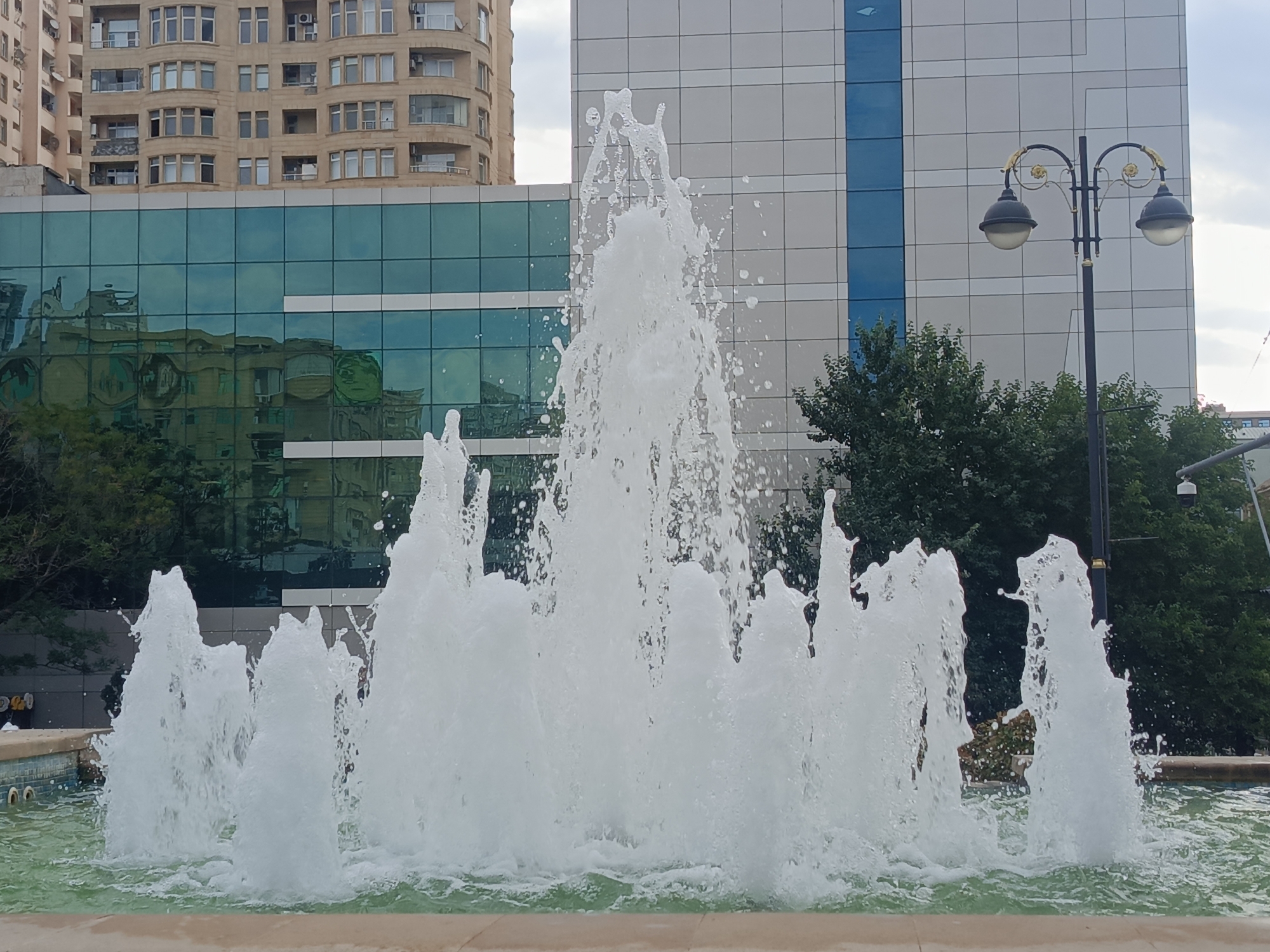The image depicts a water fountain in the foreground, with water jets of varying heights erupting from a pool. In the background, there are modern buildings with glass facades reflecting the sky and surrounding greenery. A vintage-style street lamp with two globes stands to the right of the fountain. The overall scene suggests an urban setting, likely in Baku, Azerbaijan, given the architectural style and the presence of a distinctive street lamp often found in the city. The sky is overcast, indicating a cloudy day. There are no people visible in the frame.