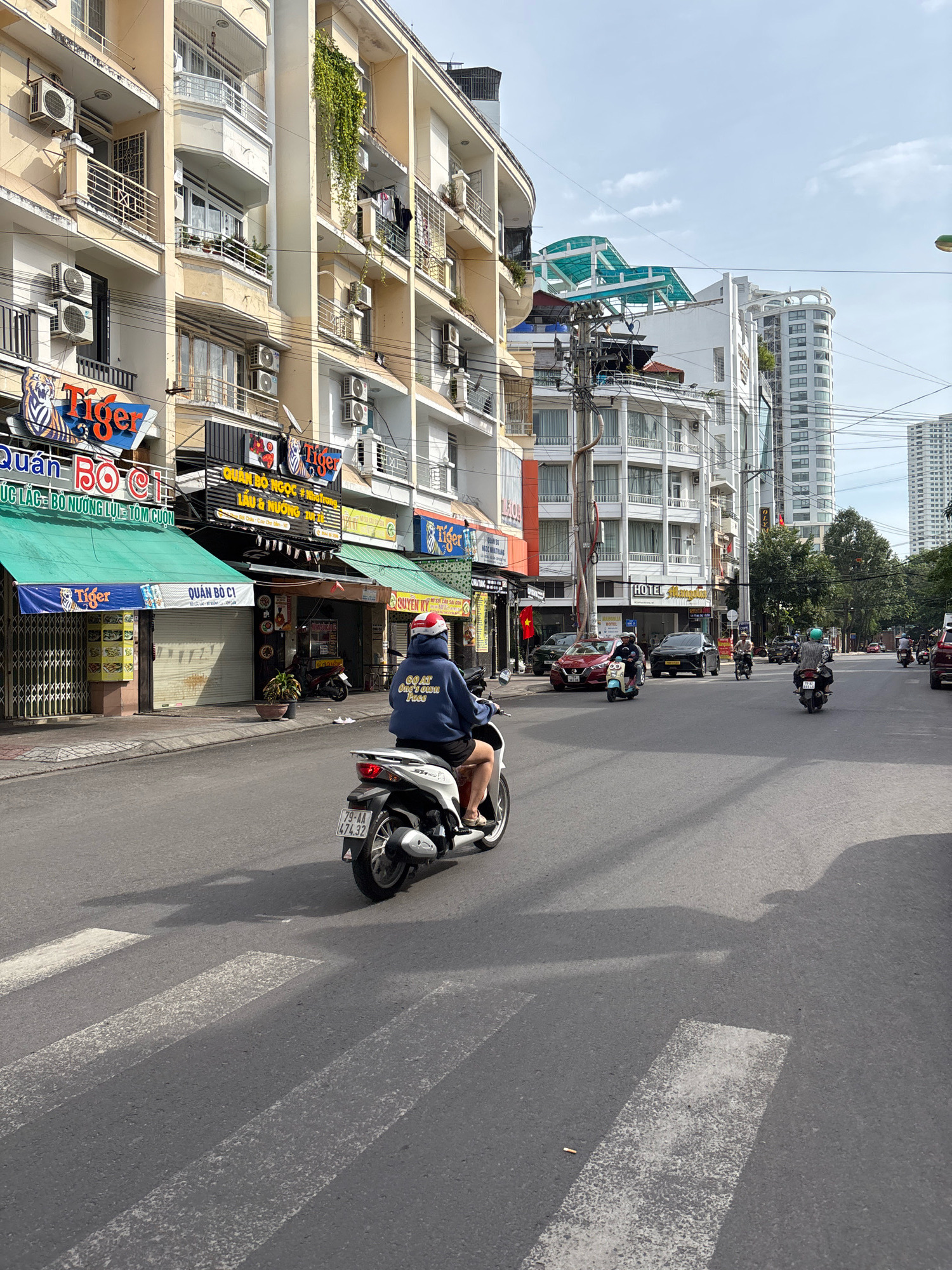 A street scene in Nha Trang, Vietnam, shows a woman riding a white scooter across a pedestrian crossing. She is wearing a dark blue hoodie, shorts, and a red and white helmet. The scooter has a license plate reading "79-AA 474.32". In the background, there are several buildings of varying heights and architectural styles, including some with balconies and air conditioning units. Businesses with signage, including "Tiger" logos, are visible on the ground floors of some buildings. The street is paved, and there are power lines and trees lining the road. The sky is partly cloudy. Other vehicles, including cars and scooters, are also present on the street, moving in the same direction as the woman.
