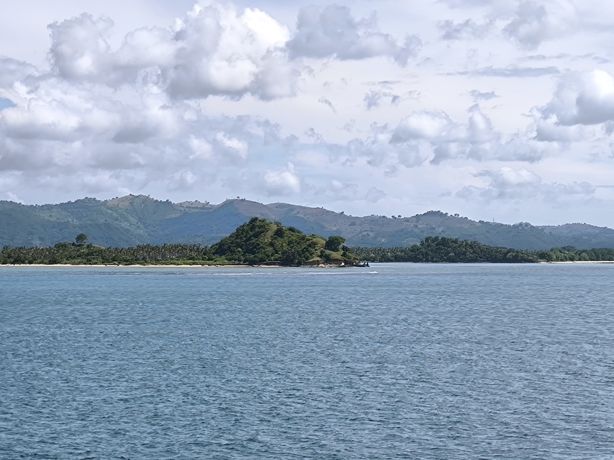 This image captures a wide-angle view of a serene seascape with islands and distant mountains under a cloudy sky.
In the foreground, a large expanse of calm, blue-green ocean water fills approximately the bottom half of the frame. The water's surface is textured with small ripples, indicating a gentle current or light breeze.
The mid-ground features a chain of green landmasses. A prominent, verdant, conical island rises from the water in the center, covered in dense foliage. To its left and right, stretches of coastline reveal narrow sandy beaches lined with abundant tropical vegetation, including numerous palm trees. A small wake can be observed in the water near the central island, suggesting the recent passage of a boat, although no vessel is clearly visible.
In the background, a range of rolling hills and mountains extends across the horizon, appearing hazy due to distance and atmospheric conditions. These hills are also covered in green vegetation, indicating a lush environment.
The sky above is largely covered by a mixture of white and grey cumulus clouds, scattered across the frame, suggesting a partly cloudy or overcast day. The lighting indicates it is daytime, likely late morning or early afternoon, with diffused light due to the clouds.
The scene depicts a natural, tranquil landscape, typical of a tropical coastal region. No people or visible text are present in the image. The location is consistent with the provided context of Lembar Selatan, Indonesia, known for its scenic islands and coastal views.