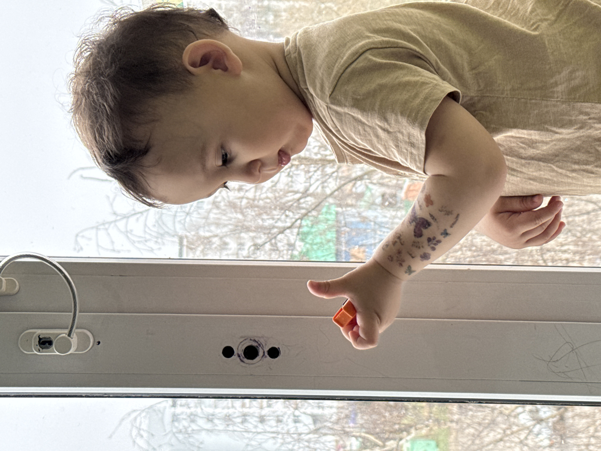 A young child, with dark hair and wearing a tan-colored shirt, stands in front of a closed white window. The child is leaning over, pointing with their right hand at the window frame. The child's arm has what appears to be temporary tattoos on it. The window has a safety latch and cable attached to it. There are three black marks on the window frame near where the child is pointing. Outside the window, bare tree branches are visible against a light sky. The city where the picture was taken is Chaikovsky, Russia.