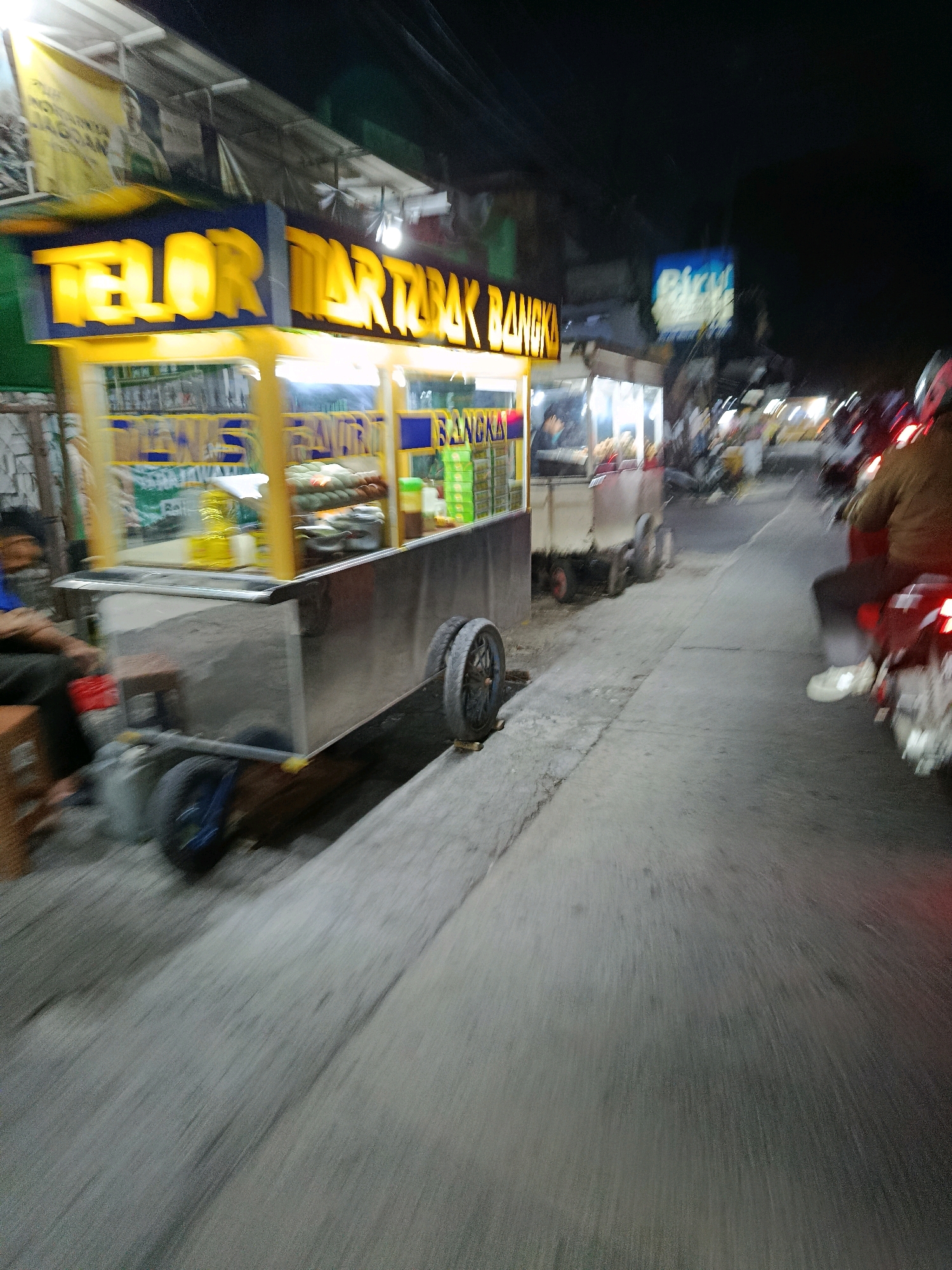 The image depicts a street scene at night in the city of Bekasi, Indonesia. In the foreground, a food cart with a bright yellow sign reading "TELOR MARTABAK" and "BANGKA" is parked on the side of the road. The cart appears to be stainless steel with wheels. Behind the cart, a person is seated on a wooden stool. The road itself is paved and a motorcyclist is visible in the middle ground, riding away from the viewer. The lighting suggests a busy street with commercial establishments. The overall image is somewhat blurry, indicating movement during capture.