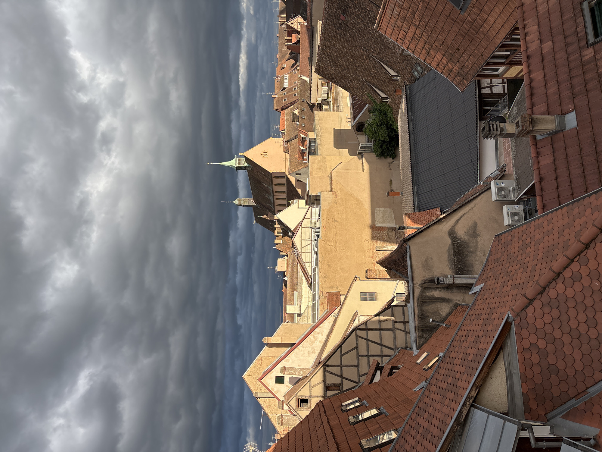This is a high-angle, medium shot of a city skyline in Colmar, France, under a dramatic, cloudy sky. The architecture is primarily composed of buildings with red tiled roofs and facades of warm, earth tones, some featuring exposed timber framing indicative of traditional Alsatian style. In the distance, a church with a green steeple rises above the surrounding rooftops. The sunlight breaks through the dark, turbulent clouds, casting strong shadows and highlighting sections of the buildings. The scene depicts a dense urban environment with a mix of residential and possibly commercial structures. No people are visible in the image.