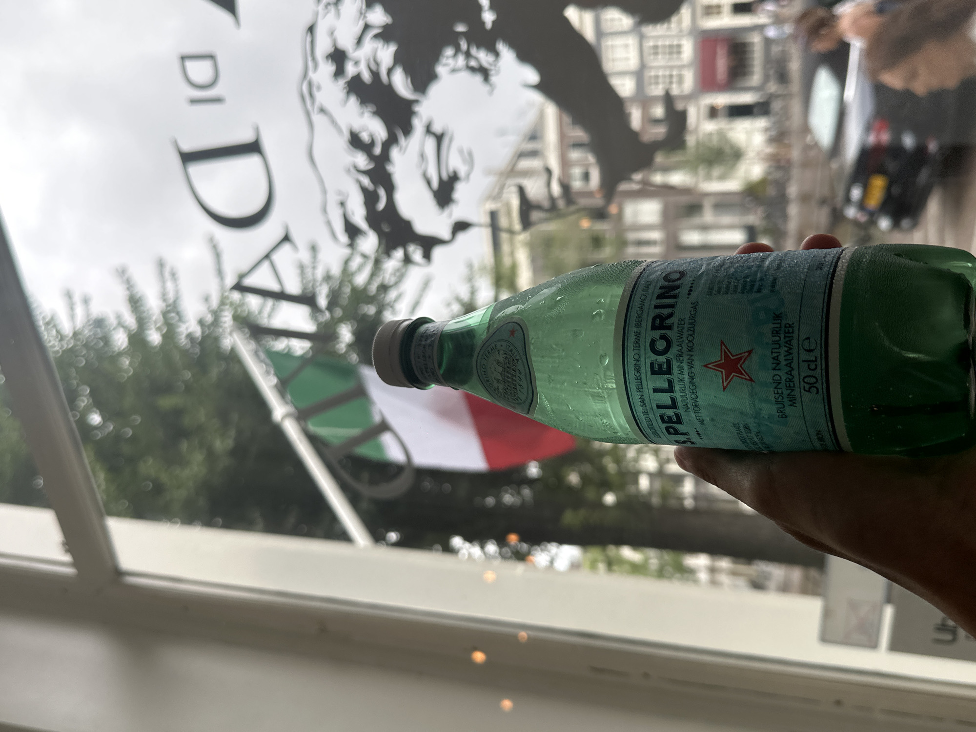 A person's hand holds a partially filled green glass bottle of San Pellegrino sparkling mineral water in front of a window. The bottle is cool and has condensation on it. The label on the bottle is light blue with white text and features a red star. Through the window, an Italian flag is visible, hanging vertically. Behind the flag and the bottle, the silhouette of a face is cast on the glass, possibly from a shadow or artwork on the window. Beyond that, a blurred cityscape of Amsterdam, Netherlands, is visible, with buildings and a street with a car and people walking. The sky appears overcast.
