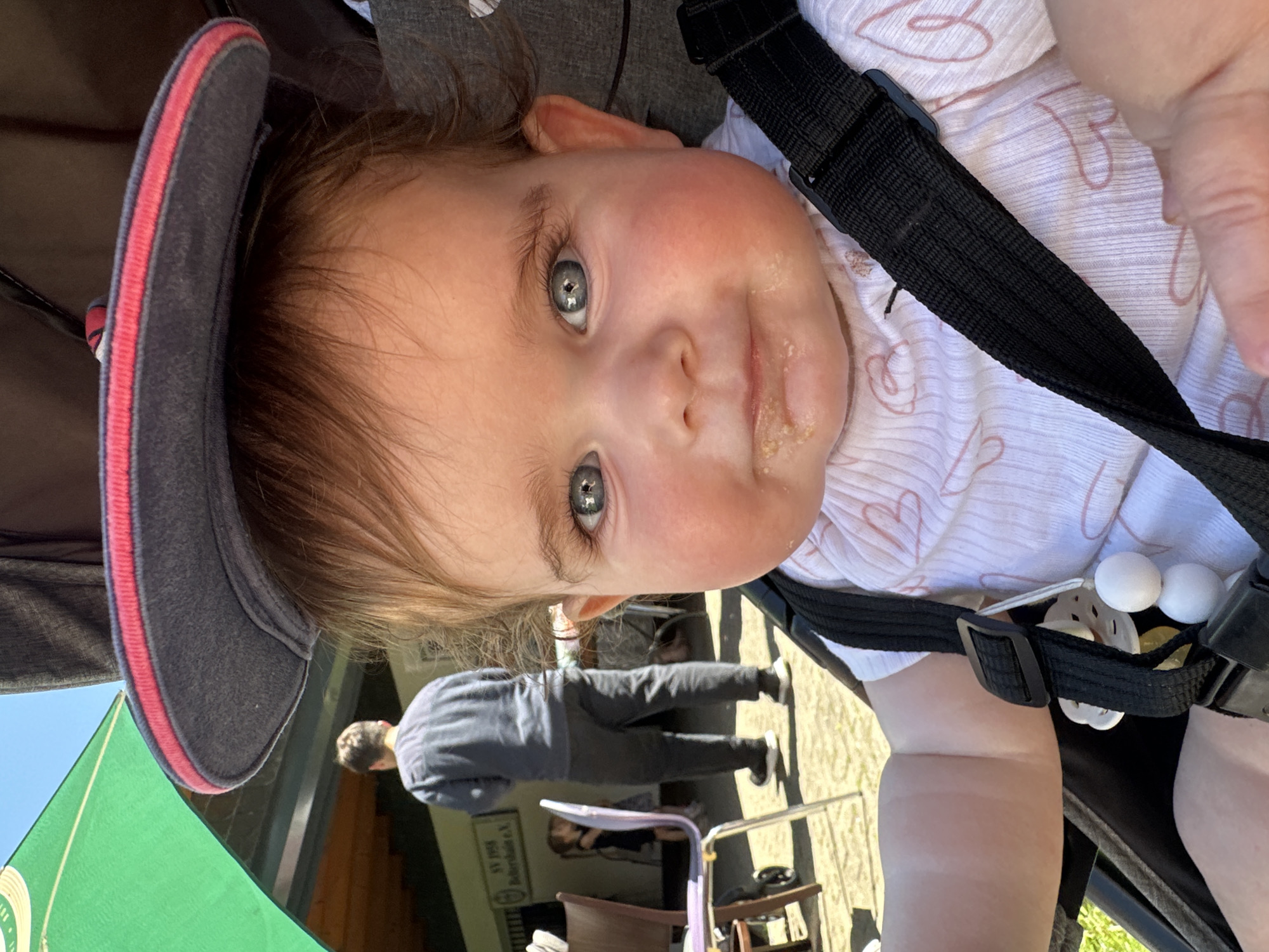 A baby with blue-gray eyes is strapped into a stroller with a gray sun visor trimmed with red. The baby has dribble on its face. A green banner with white text and an emblem is to the left of the stroller. A man in a gray hooded sweatshirt is walking on a paved area behind the stroller. A sign with the text "SV 1925" and "Bieberdale e.V." is visible on a building in the background. The location is most likely in the city of Lumda, Germany.
