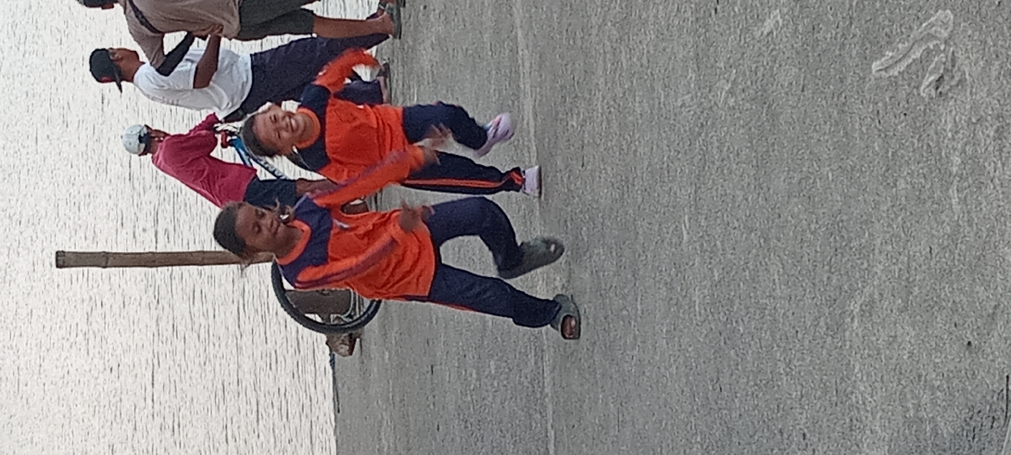 Two young girls in orange and navy athletic wear are running on a paved surface near a body of water. The girl on the left is wearing dark slip-on shoes and the girl on the right is wearing pink athletic shoes. Behind them, a bicycle with a blue frame is visible, along with several adults. One adult is wearing a white t-shirt and a black baseball cap, and another is wearing a red long-sleeved shirt. A wooden post stands in the background on the left. The location is identified as Kalipucang, Indonesia.