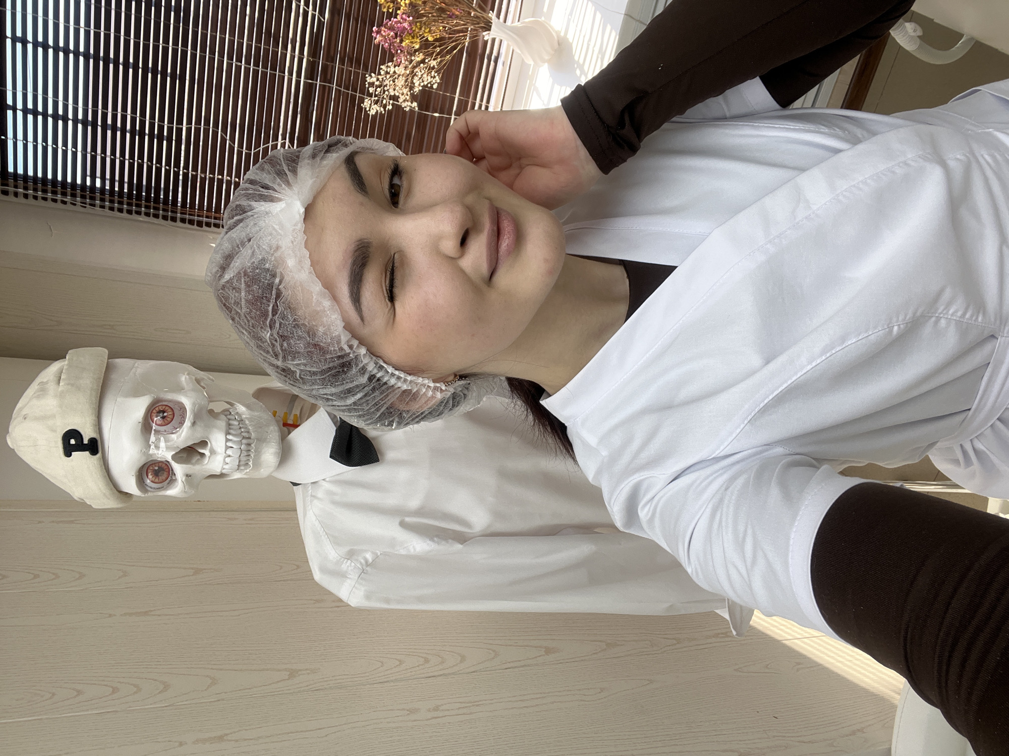 A young woman in a white lab coat and hairnet is posing for a selfie with a skeleton. The skeleton is wearing a white hat with a "P" on it, a white shirt, and a black bow tie. They are standing in front of a light-colored wall, with a window and a vase of flowers visible in the background. The location is likely indoors, but the exact location is unknown, and it is not possible to confirm if it is in Neftekamsk, Russia, without additional information.