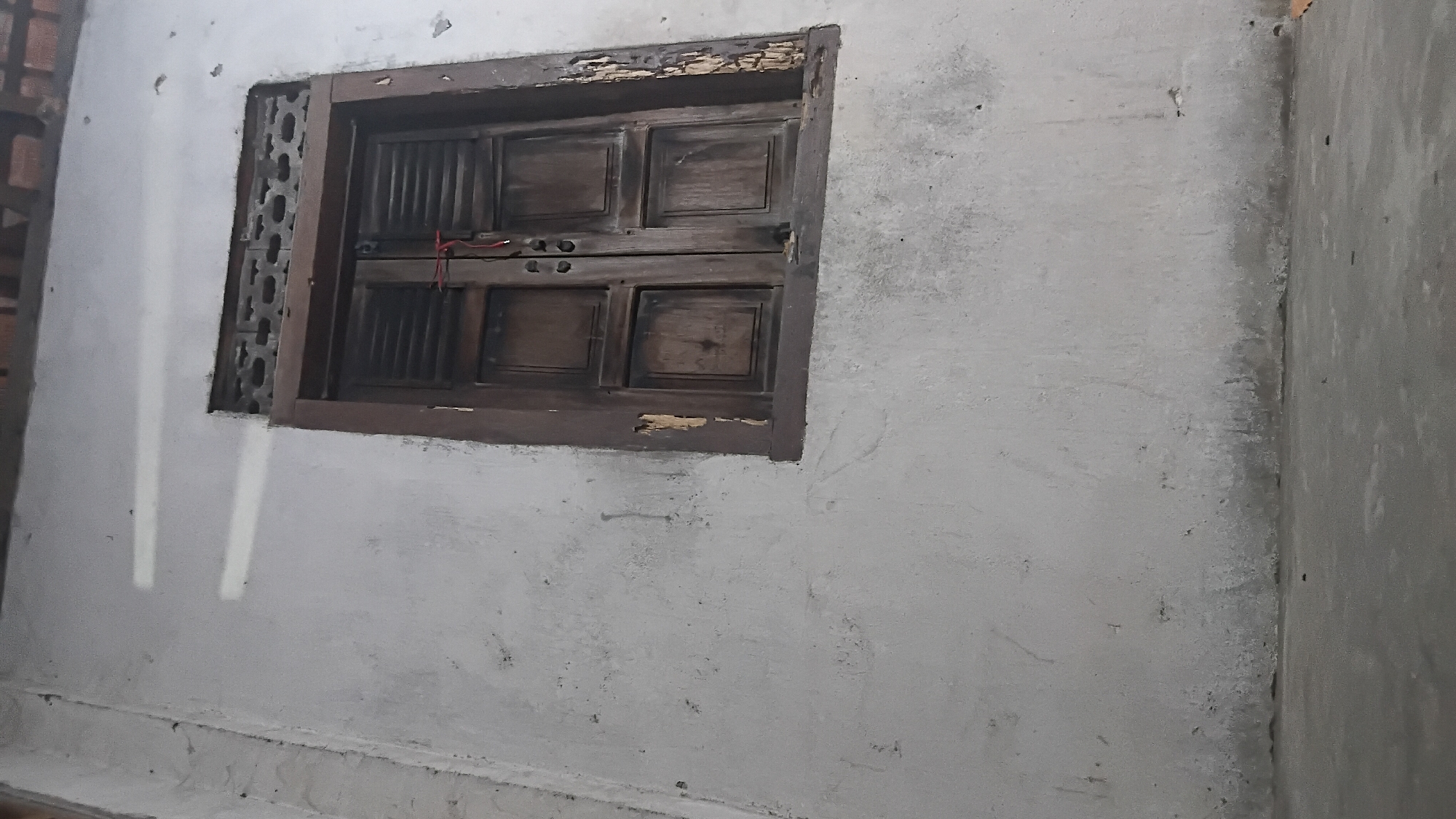 The image shows a close-up of a weathered wooden window set into a textured, off-white wall. The window has two wooden shutters, each divided into two panels, with visible louvers. A thin red string is tied around the handles of the shutters, suggesting they are secured. The wooden frame of the window is dark brown and shows signs of decay and peeling paint, particularly on the right side. Above the shutters, a decorative vent with a patterned grill is visible. The wall itself appears old, with a rough, uneven surface and some staining or discoloration, especially towards the bottom. The ground in front of the wall is a concrete surface. The lighting in the scene creates bright, elongated shadows on the wall from an unseen light source. The overall impression is one of an aged and somewhat neglected structure, possibly in a residential area of Prabumulih, Indonesia, given the context. There are no people or animals visible in the image.