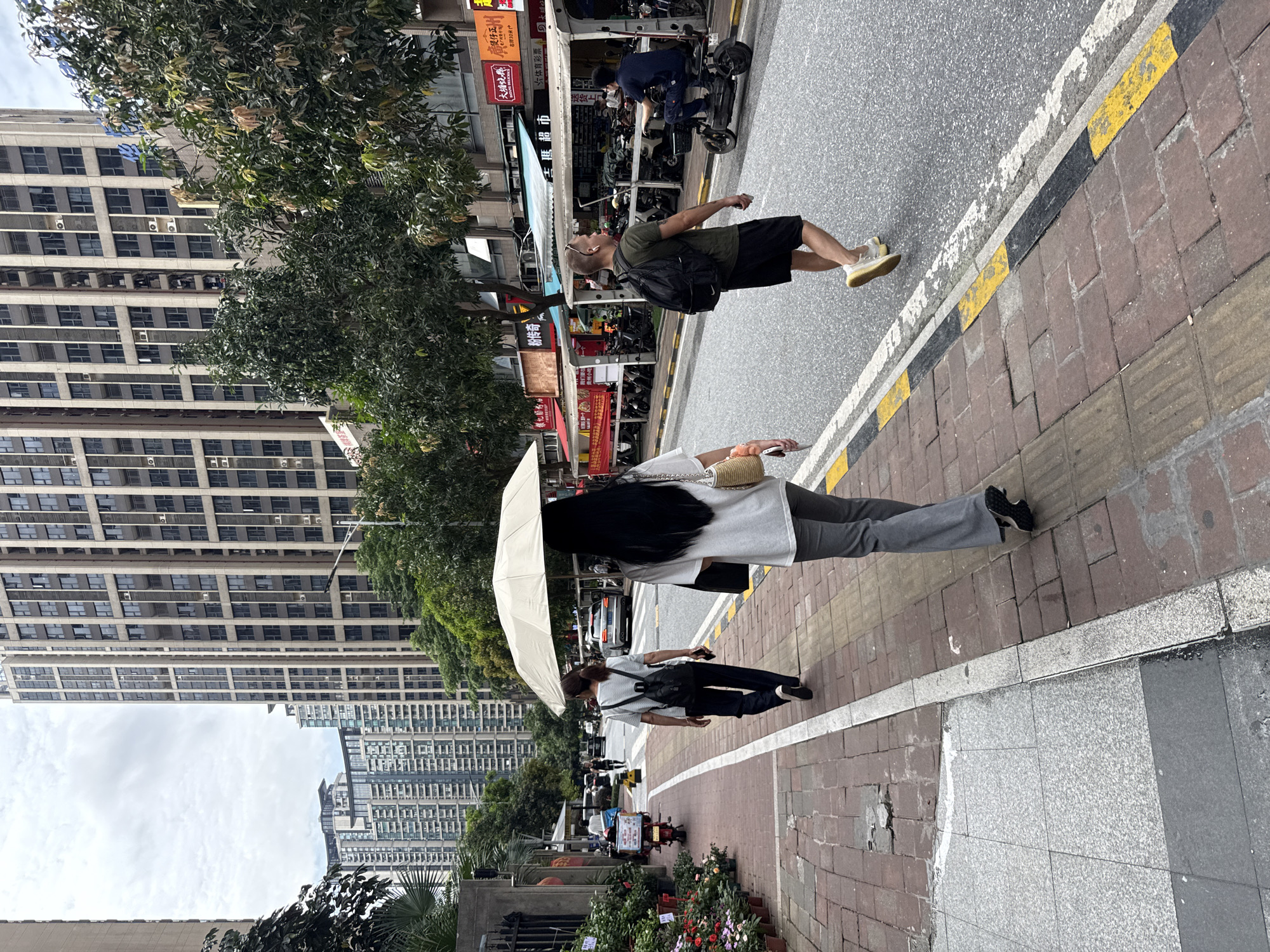 A street scene in Haizhu District, China, features a sidewalk with brick paving and a road. Two women are walking on the sidewalk, one holding a large white umbrella. A man with a backpack is walking on the road, and another person is riding a three-wheeled vehicle. Several buildings, including tall high-rise apartments, are visible in the background. Trees line the street, and various storefronts with awnings are present. The sky is overcast.