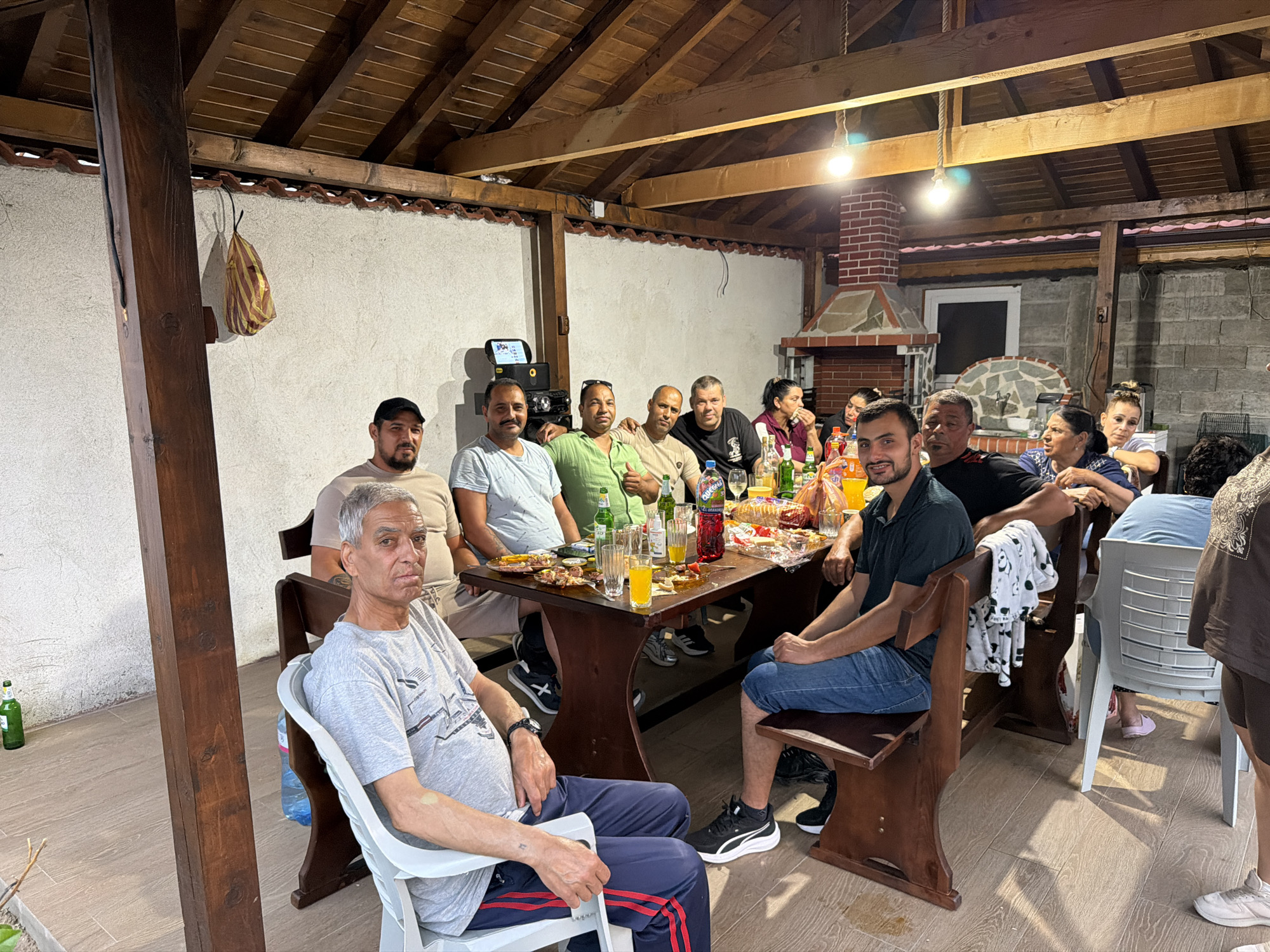 An outdoor social gathering of approximately fifteen to twenty individuals, primarily men, is captured under a wooden-beamed pergola structure in Sinitovo, Bulgaria. The group is seated around a large, dark wooden table heavily laden with food items, including bread, various snacks, and numerous bottles of beverages such as soda, beer, and water, along with glasses containing orange liquid. Most attendees are adults dressed in casual clothing. A gray-haired man in a light gray t-shirt occupies a white plastic chair in the immediate foreground, looking directly at the camera. To the right, another man in a dark green polo shirt and jeans is also looking towards the camera. A man in a light green shirt, seated further down the table, offers a thumbs-up gesture. In the background, a prominent brick barbecue and chimney structure is visible on the right, beside a window and a concrete block wall. A portable speaker or audio device rests on a smaller stand to the left of the main table. A decorative, wrapped item hangs from a wooden beam near the left. The floor consists of light-colored tiles, and the area is illuminated by several hanging light bulbs. A small green beer bottle is visible on the ground to the far left.