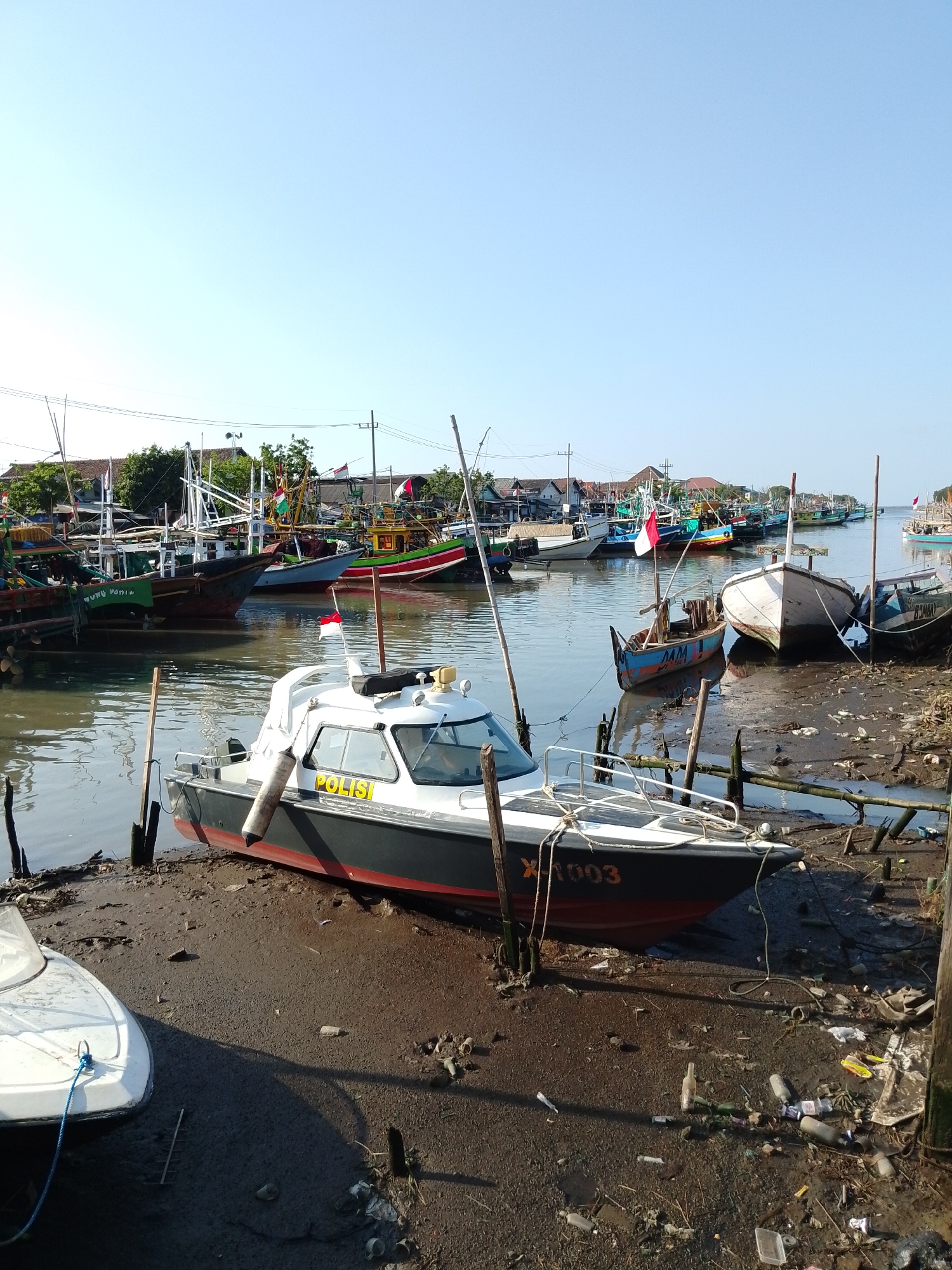 The image depicts a harbor scene in Pasuruan, Indonesia, likely on the coast or along a river, characterized by a collection of various boats. In the foreground, a white and dark-hulled police boat with the word "POLISI" visible on its side is partially grounded on a muddy bank. The boat is equipped with a cabin and is tied to shore with ropes. To the left of the police boat, another smaller white boat is also partially visible, also grounded on the mud.
The harbor is filled with numerous other fishing boats of varying sizes and designs, some of which are moored in the water, while others are also grounded or appear to be docked. Many of these boats have masts and rigging, suggesting their use for fishing. Several Indonesian flags are visible on some of the boats, indicating national affiliation. The water appears murky, and the shoreline is muddy and littered with debris, including plastic bottles and other waste.
In the background, there is a line of buildings and houses that appear to be part of a coastal or riverside settlement. Trees and vegetation are also present. The sky is clear and bright blue, suggesting a sunny day, and the overall lighting indicates it is likely morning or late afternoon. The scene suggests a working harbor, possibly a commercial fishing port or a maritime police outpost, with elements of everyday life and the presence of both operational vessels and those possibly undergoing maintenance or docked for the day.