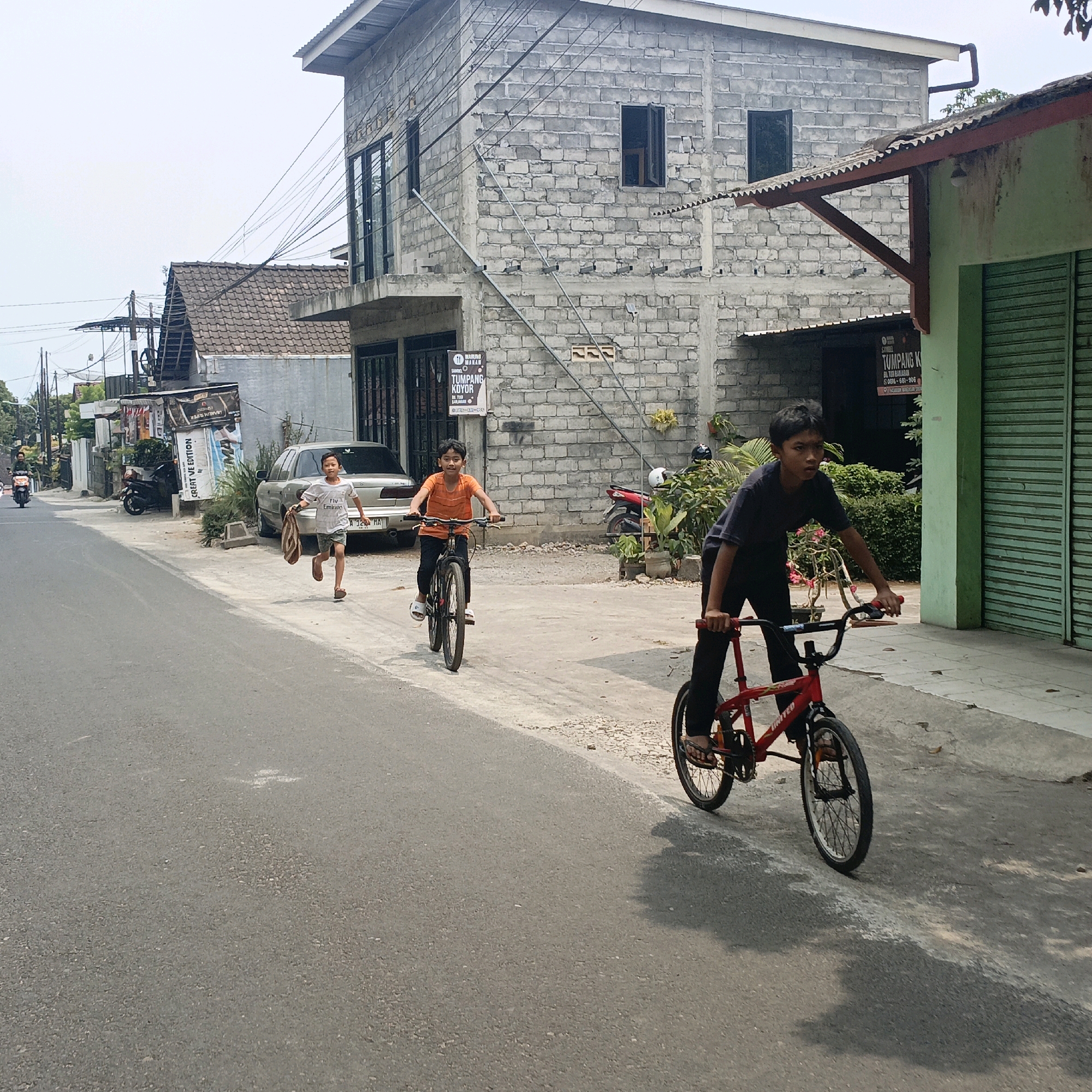 The image depicts a street scene in Salatiga, Indonesia, on a bright, overcast day. Three young boys are the central focus. In the foreground, a boy wearing a dark t-shirt and black pants rides a red BMX bicycle. His right hand is on the handlebars, and his left hand is positioned near the brake. Behind him, another boy in an orange t-shirt and black pants rides a black bicycle, looking ahead. To the left, a third boy with dark hair runs alongside the road, carrying a light-colored bag over his right shoulder. He wears a white t-shirt with the text "Fly Emirates" and shorts.
The street is paved with asphalt. On the right side of the street, a light green building with a corrugated metal roof and green metal roll-up doors is visible. A red-painted wooden bracket supports the roof overhang. Adjacent to this building, a multi-story structure constructed of concrete blocks with a metal roof is under construction or recently completed, featuring large dark-framed windows. A sign with Indonesian text, including "TUMPANG KOYOR," is attached to the block wall.
On the left side of the street, a silver sedan is parked, and further down, a black motorcycle is also parked. A sign with "CREATIVE EDITION" printed on it stands near the parked vehicles. In the distance, more houses and trees line the road, and a person on a motorcycle travels away from the camera. Overhead, a dense network of power lines crisscrosses the sky. The overall environment suggests a residential or commercial street in a built-up area.