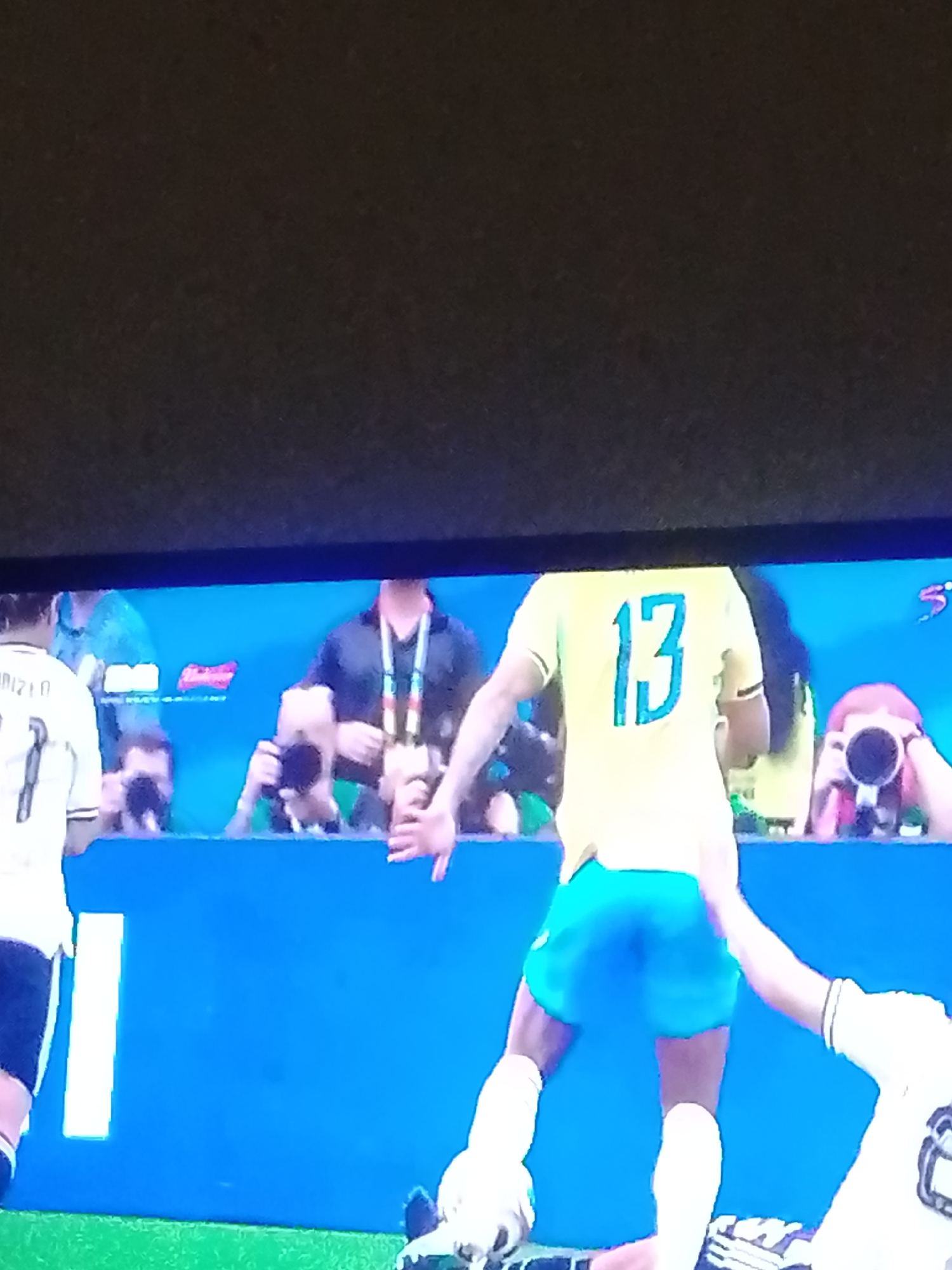 The image shows a close-up view of a soccer game with several individuals visible. In the foreground, a player wearing a yellow jersey with the number 13 on the back is the central focus. Their back is to the camera, and they are wearing blue shorts. To the left, another player is partially visible, wearing a white jersey with a number 1 on it and black shorts. In the background, several other people are standing behind a blue barrier, some of whom appear to be photographers holding cameras. The environment looks like a sports stadium or arena with a bright blue backdrop and green turf visible at the bottom of the image. The lighting suggests an indoor or evening event.