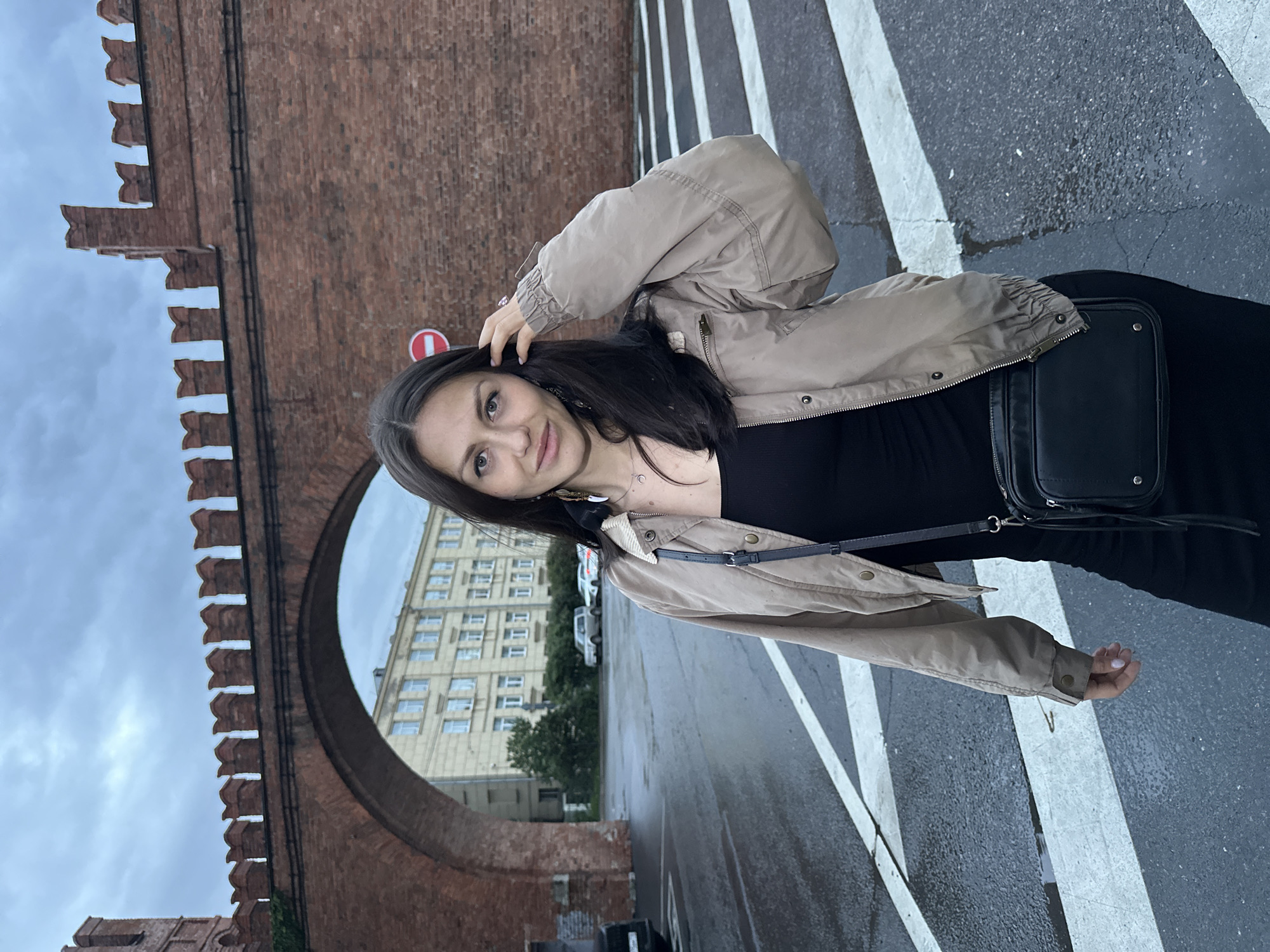 The image captures a woman standing on a wet crosswalk in Saint Petersburg, Russia. She is positioned in front of a historic brick structure with an arched gateway and crenellated battlements. Behind the archway, a light-colored, multi-story building with numerous windows is visible, along with parked cars. The sky is overcast, suggesting a cloudy day. The woman is wearing a black dress, a beige bomber jacket, and a black crossbody bag. She is touching her hair with her right hand, and her gaze is directed towards the camera. The crosswalk markings are stark white against the dark, wet asphalt.