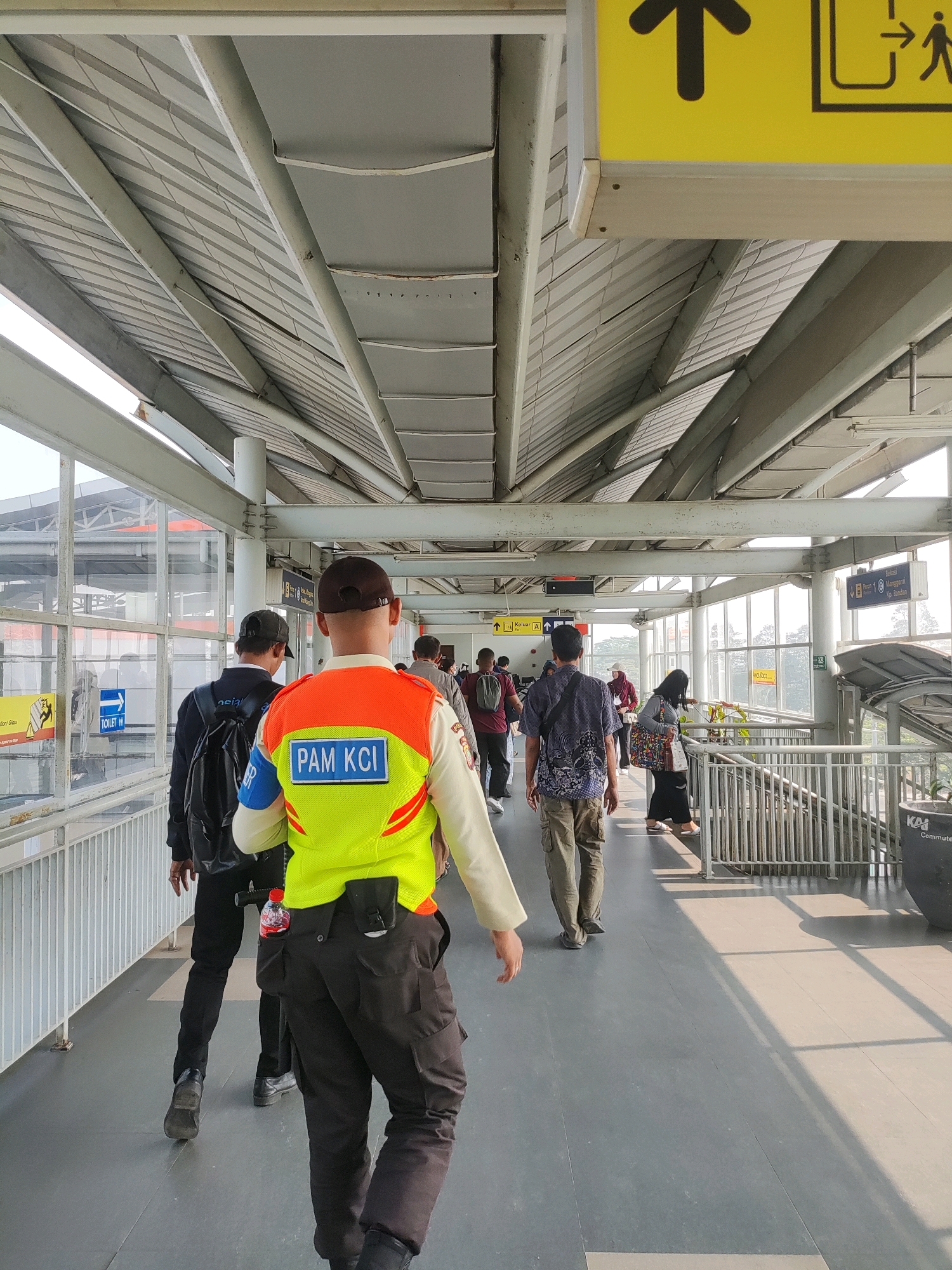 The image depicts a covered walkway at a train station in Telagamurni, Indonesia. In the foreground, a person wearing a bright yellow and orange vest with "PAM KCI" written on it walks away from the camera. They are also wearing dark pants and a baseball cap. To their left, another person in a dark jacket and baseball cap walks ahead, carrying a backpack. Several other individuals are visible further down the walkway, some walking and others standing. The walkway has a metal roof supported by pillars, with large windows along the side offering a view of the exterior. Signage is present, including directional arrows and an icon indicating a toilet. The ground is paved with large, grey tiles. Sunlight streams through the windows, creating strong shadows on the walkway.