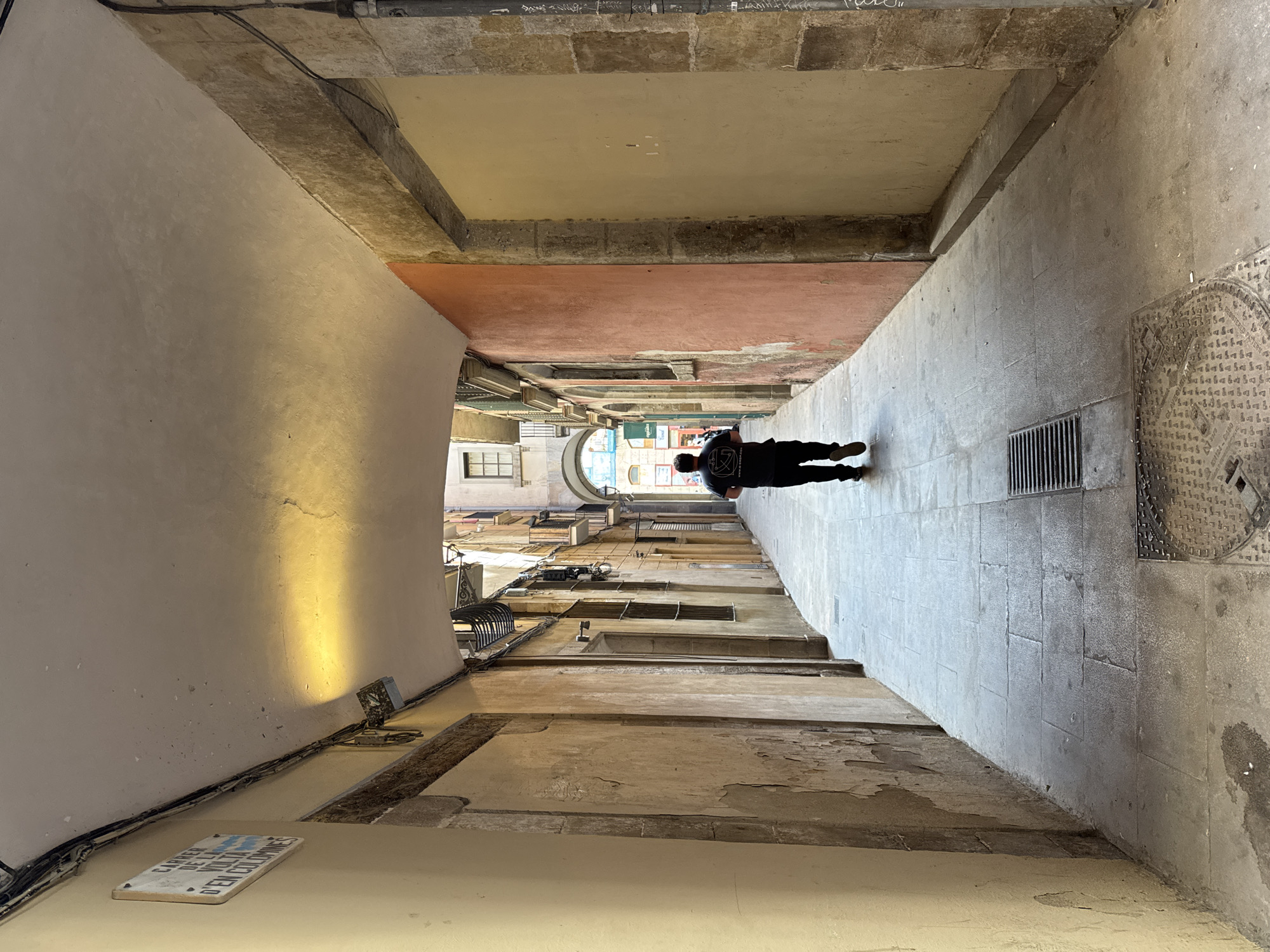 The image shows a narrow, stone-paved street in Barcelona, Spain. The perspective is shot looking down the length of the street with a man standing roughly in the middle of the frame. He is wearing a black t-shirt and appears to be walking away. The buildings lining the street are old with weathered facades and windows. The street is covered by a series of archways. A sign on the wall reads "Carrer de la Volta d'En Colomines,". A manhole cover can be seen on the right. The light is muted but brightens towards the end of the street revealing a fragment of the city's architecture.