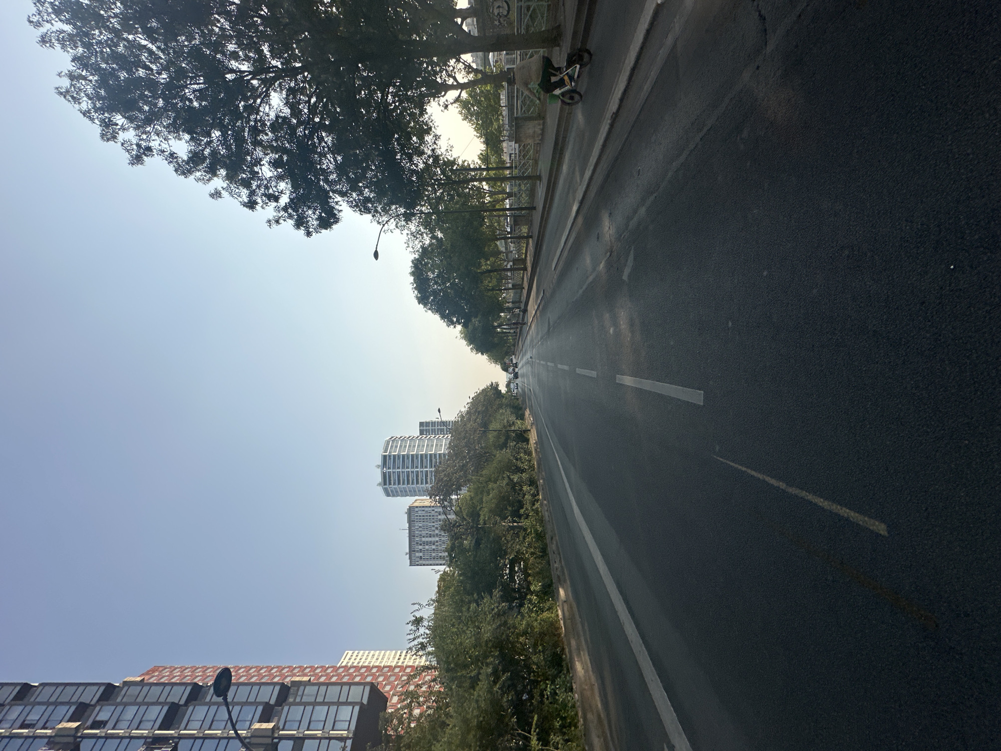 The image shows a street scene in Paris, France, likely in the late afternoon or early evening based on the lighting. The perspective is from a low angle, looking down a wide, empty road that curves gently to the left in the distance.
On the left side of the road, there is a tall, modern building with a red and glass facade, followed by a row of trees and then several taller, white apartment buildings with distinctive rounded tops. The road itself has white lane markings.
On the right side of the road, there is a sidewalk lined with trees. A person is riding an electric scooter or bicycle along the right side of the road, on what appears to be a dedicated path or the edge of the roadway. Beyond the trees and sidewalk, there's a metal fence and then some low-lying structures or walls. The sky is clear and pale blue, suggesting fair weather. The overall impression is one of an urban environment with a blend of modern architecture and mature trees.