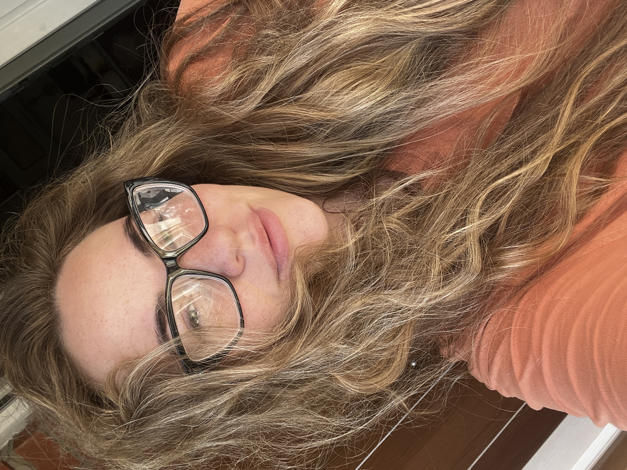 A close-up, overhead selfie shows a young Caucasian woman with long, blonde, wavy hair and fair skin with freckles. She is wearing black-rimmed glasses and an orange t-shirt. Her head is tilted to the left and her hair is spread around her face and shoulders. The background is blurred, but appears to be outdoors, with wooden planks on the left and a dark opening on the right. The image does not contain information to determine its location in Chaikovsky, Russia.
